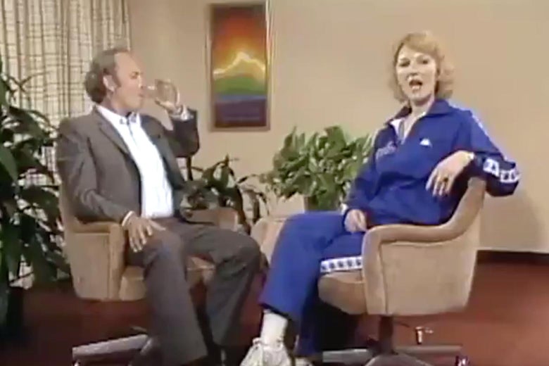 Leta Powell Blake sits across from a man drinking from a glass as she addresses the camera during an interview on a '70s-era TV set