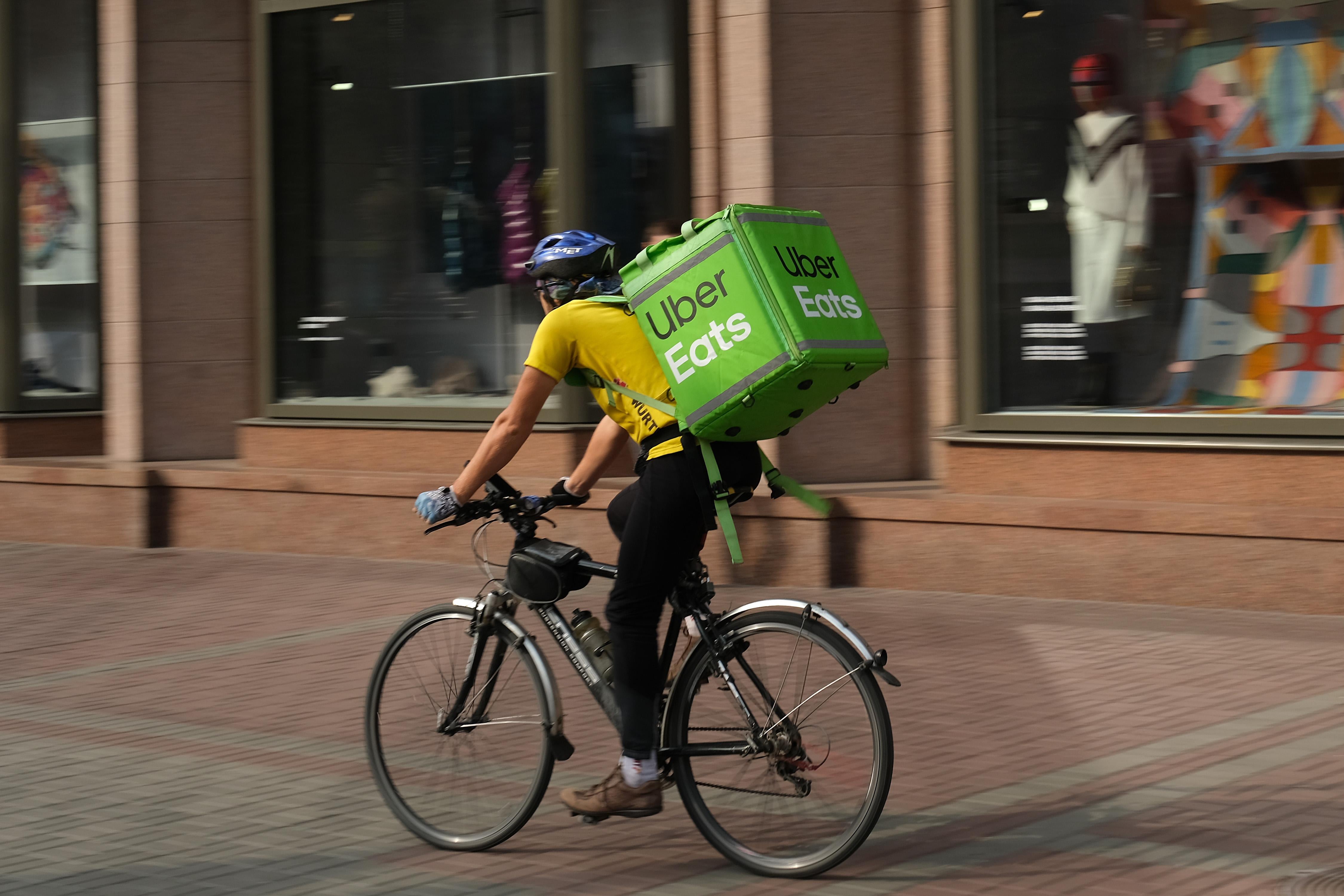 A man wearing a yellow shirt rides a bike past storefronts. On his back is a green Uber Eats container.