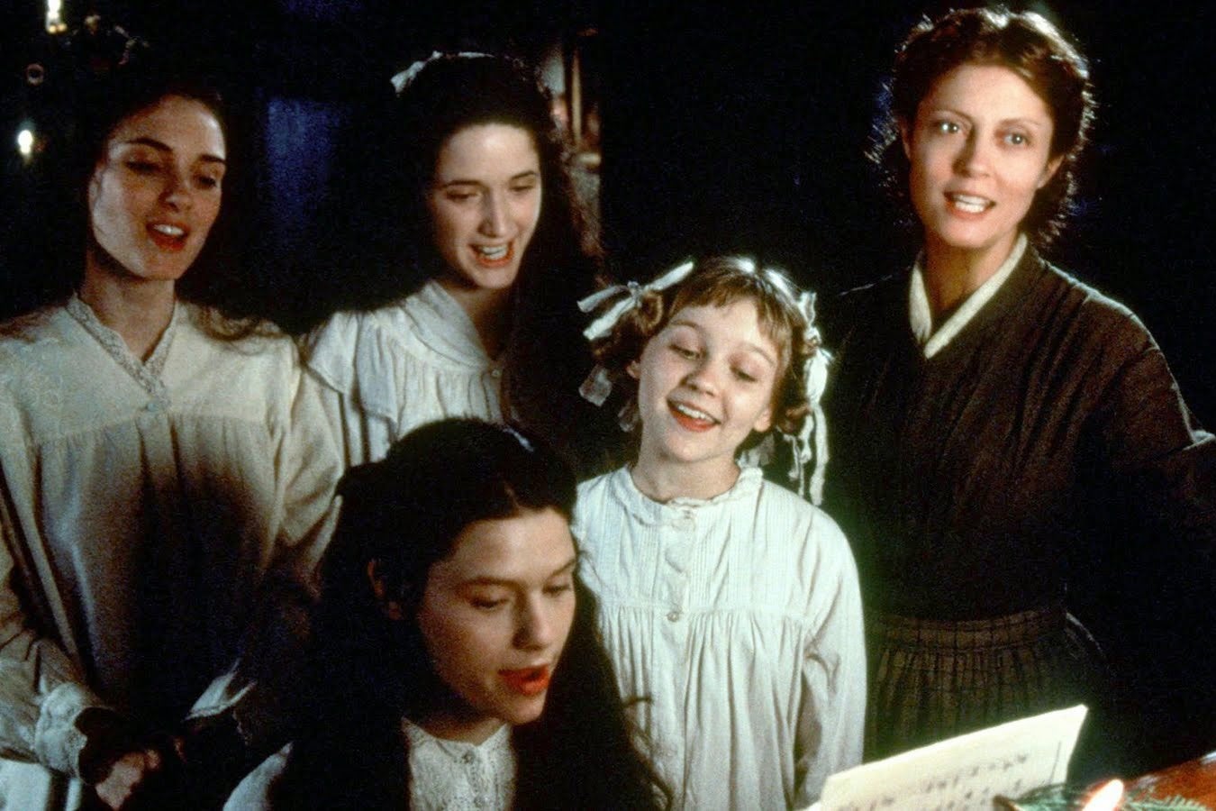 A young girl with long black hair dressed in a white dress plays the piano while three young women also dressed in white stand behind her singing along. Their mother dressed in brown stands by their side behind the piano also singing along.