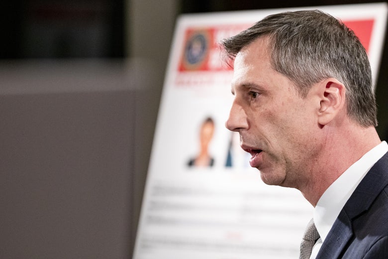 A man speaks with a blurry Wanted poster behind him.