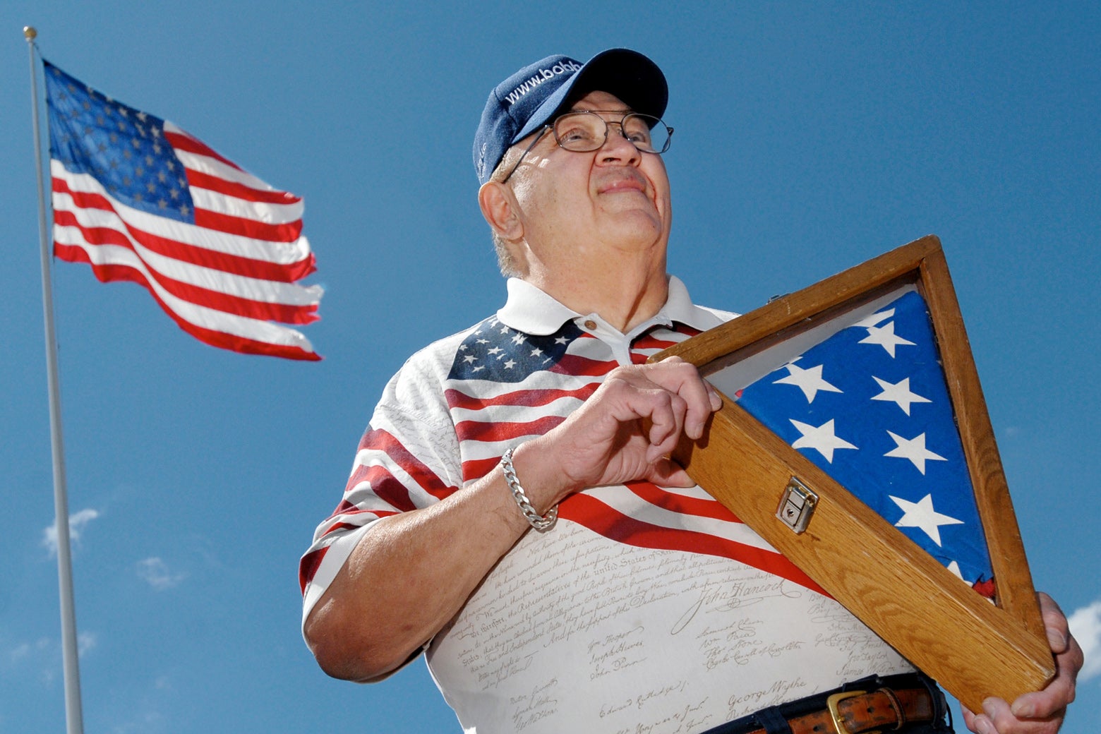 A man in a flag polo shirt holds up a flag in a wooden box. A flag also flies behind him against the blue sky.