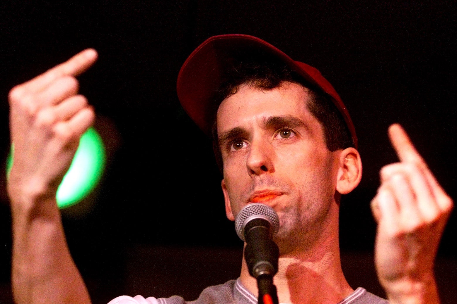 Dan Savage, wearing a red baseball cap and behind a microphone, points both index fingers updward.