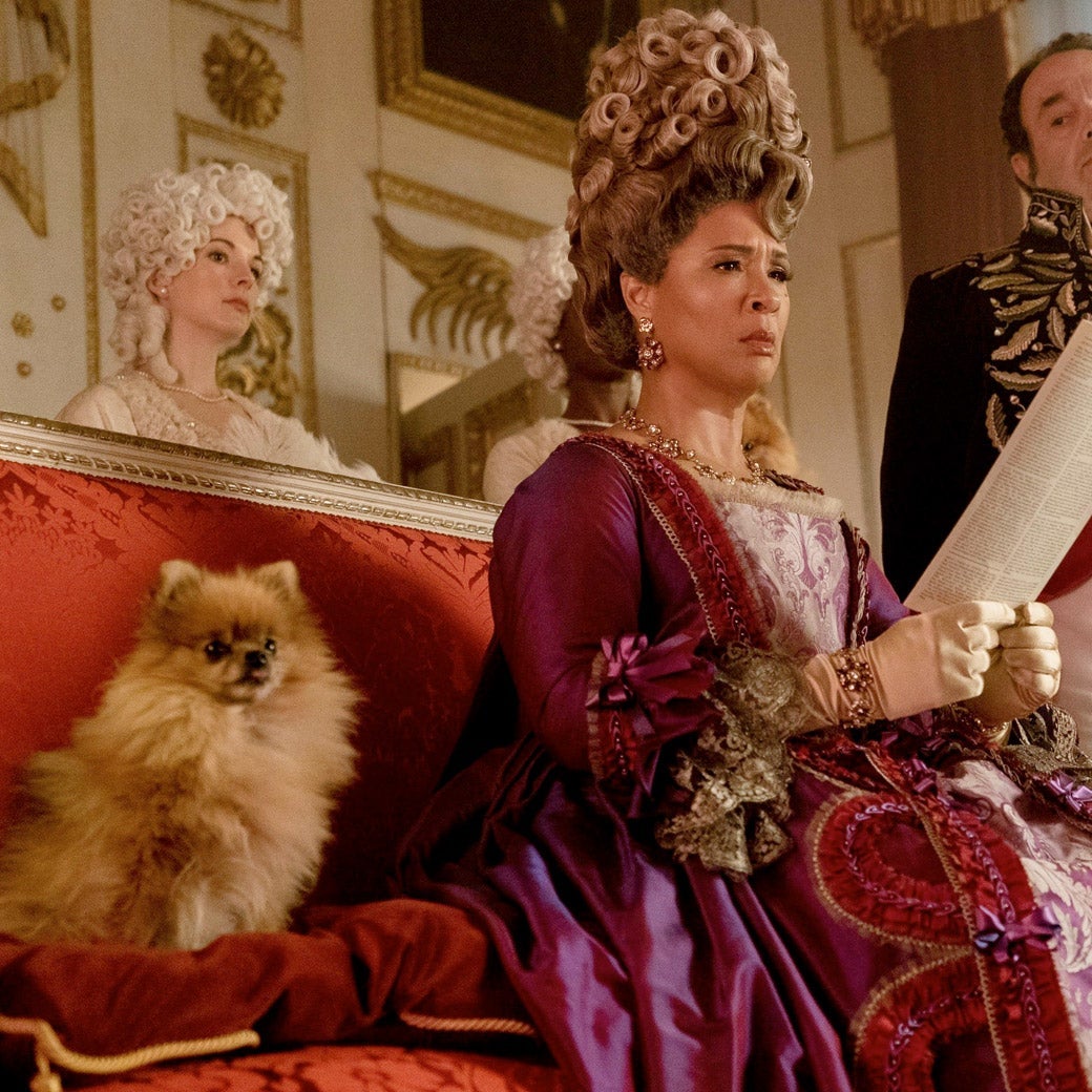 Golda Rosheuvel as Queen Charlotte in Bridgerton, sitting on a couch and positioned between a dog and a standing man