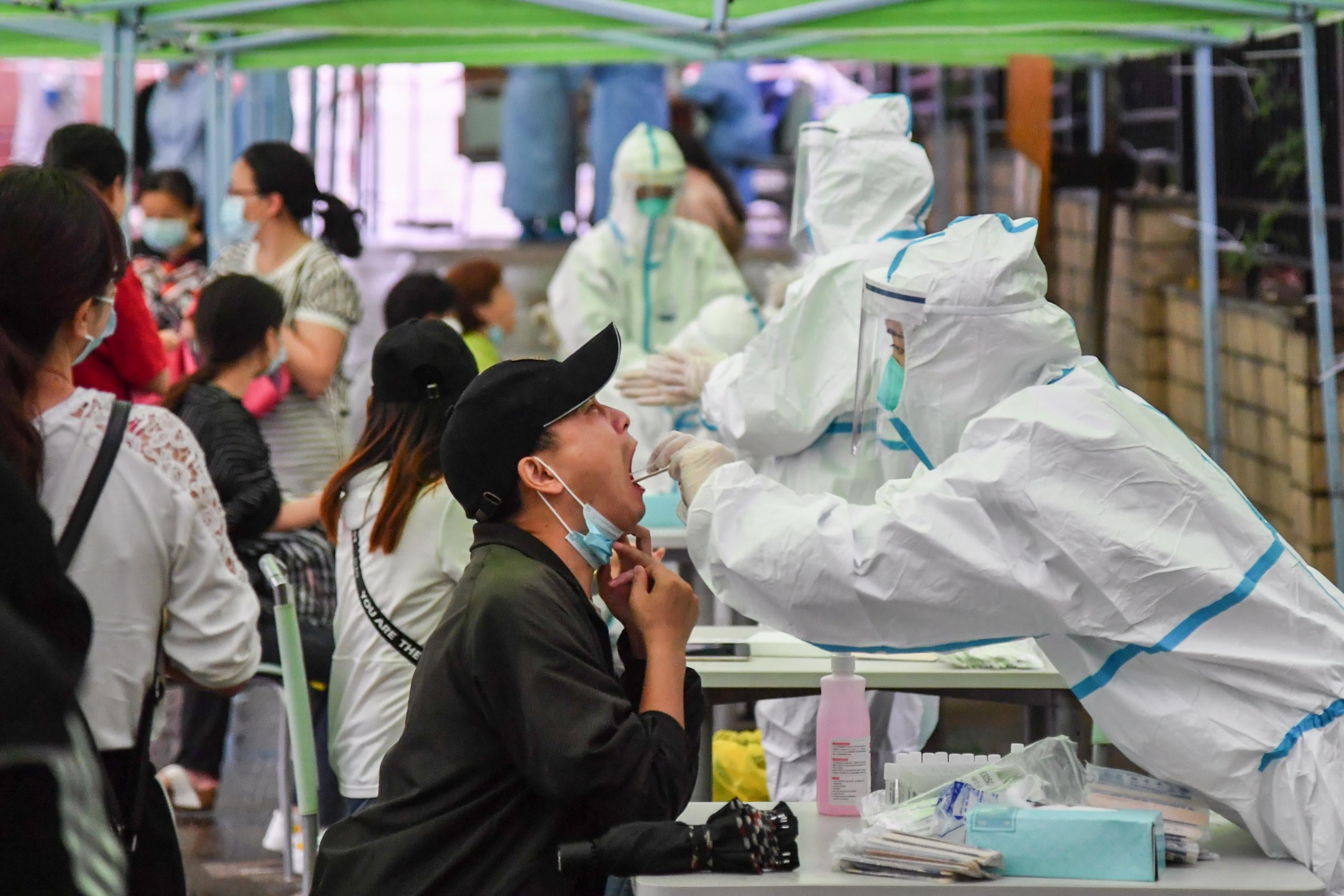 A man, sitting down, opens his mouth wide as a medical worker in personal protective equipment takes a swab sample from his throat. Others wait in line behind the man. 
