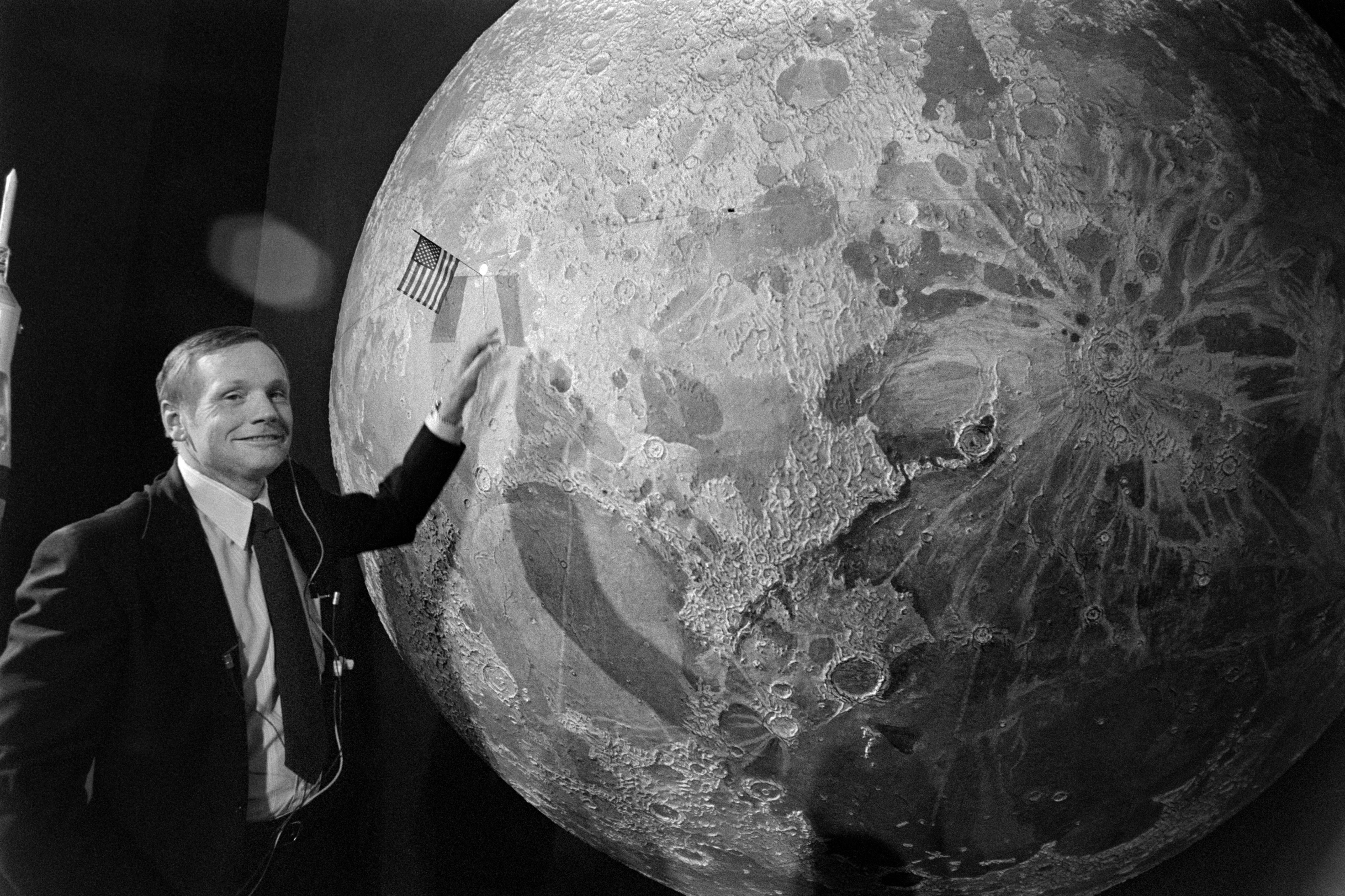 Neil Armstrong poses next to a large picture of the moon that has a tiny U.S. flag sticking out of it.