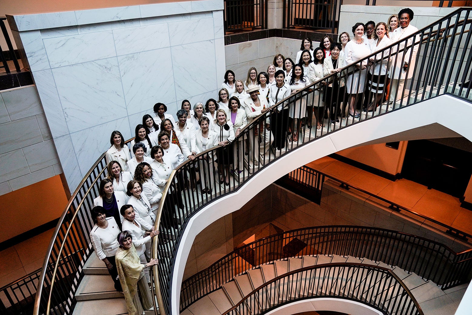 Nancy Pelosi and congresswomen wearing white pose for a photo on a spiral staircase.