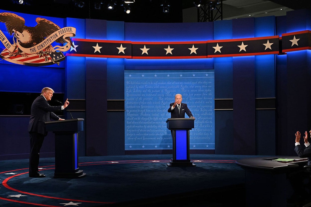 Biden, seen from a distance, gestures directly toward the camera while Trump, to the left of the frame, gestures toward the moderator's table.