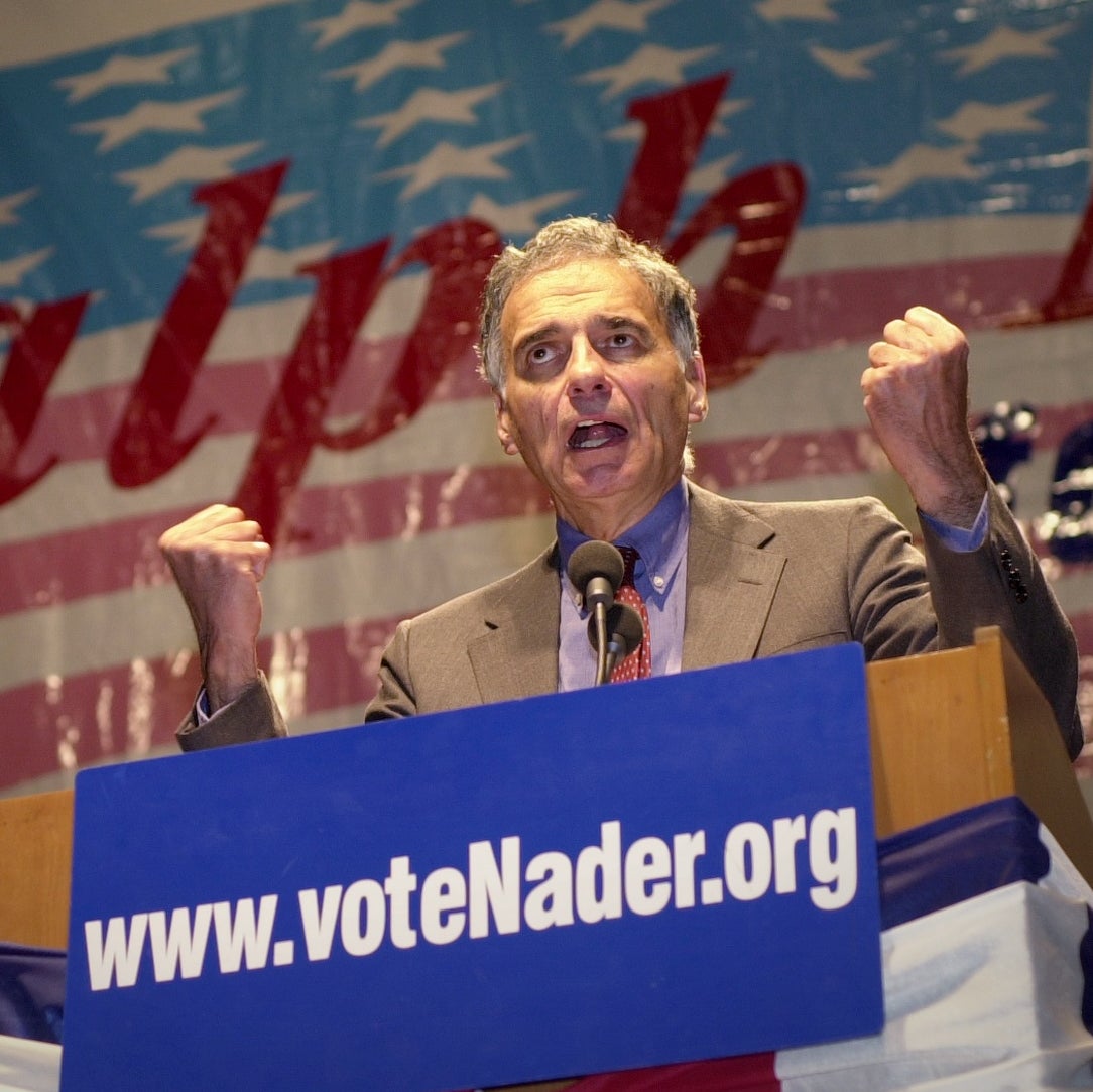 Nader speaks at a podium with both fists clenched in front of him, an American flag with his name one it behind him, and a sign on the podium reading www.voteNader.org.