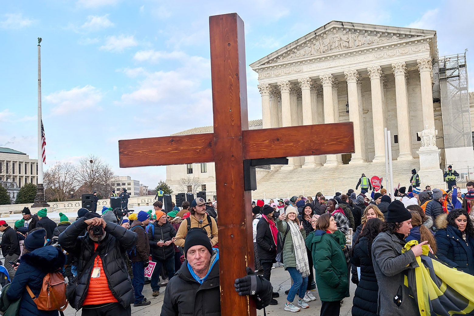 A man holds a larger wooden cross within a crowd in front of the Supreme Court building.