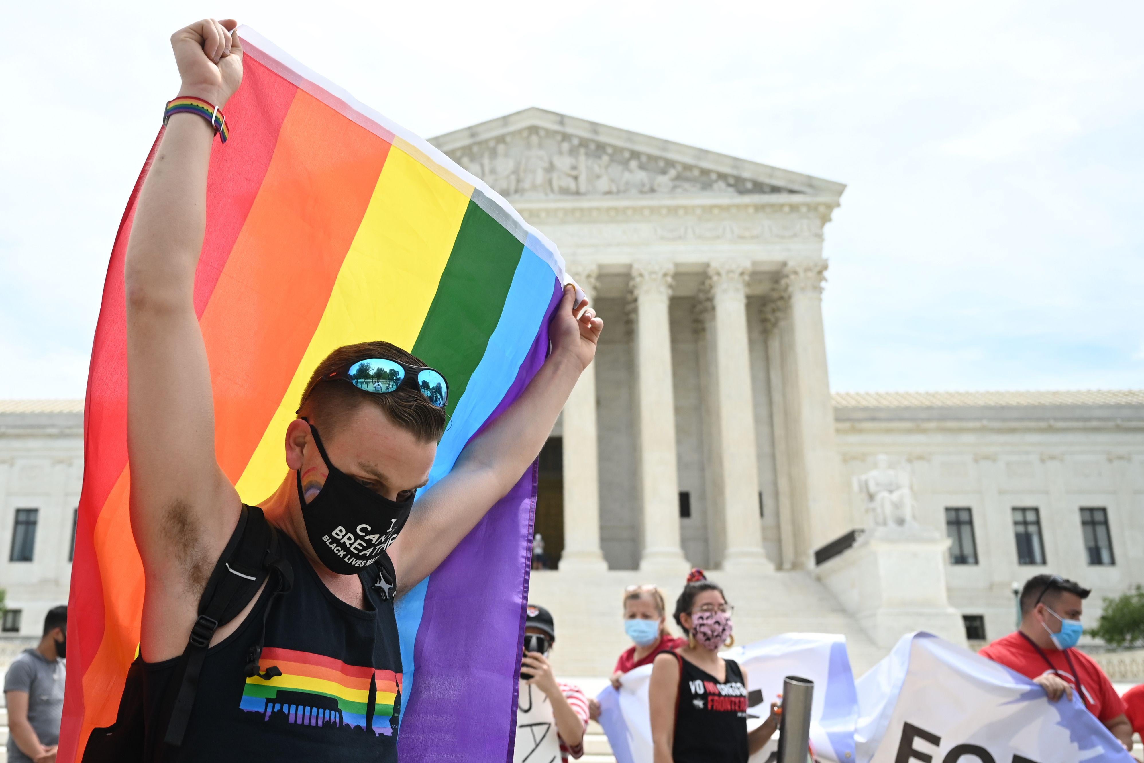 Man wearing a black mask and shirt with sunglasses on his head drapes holds a rainbow flag above his head in front of the Supreme Court.