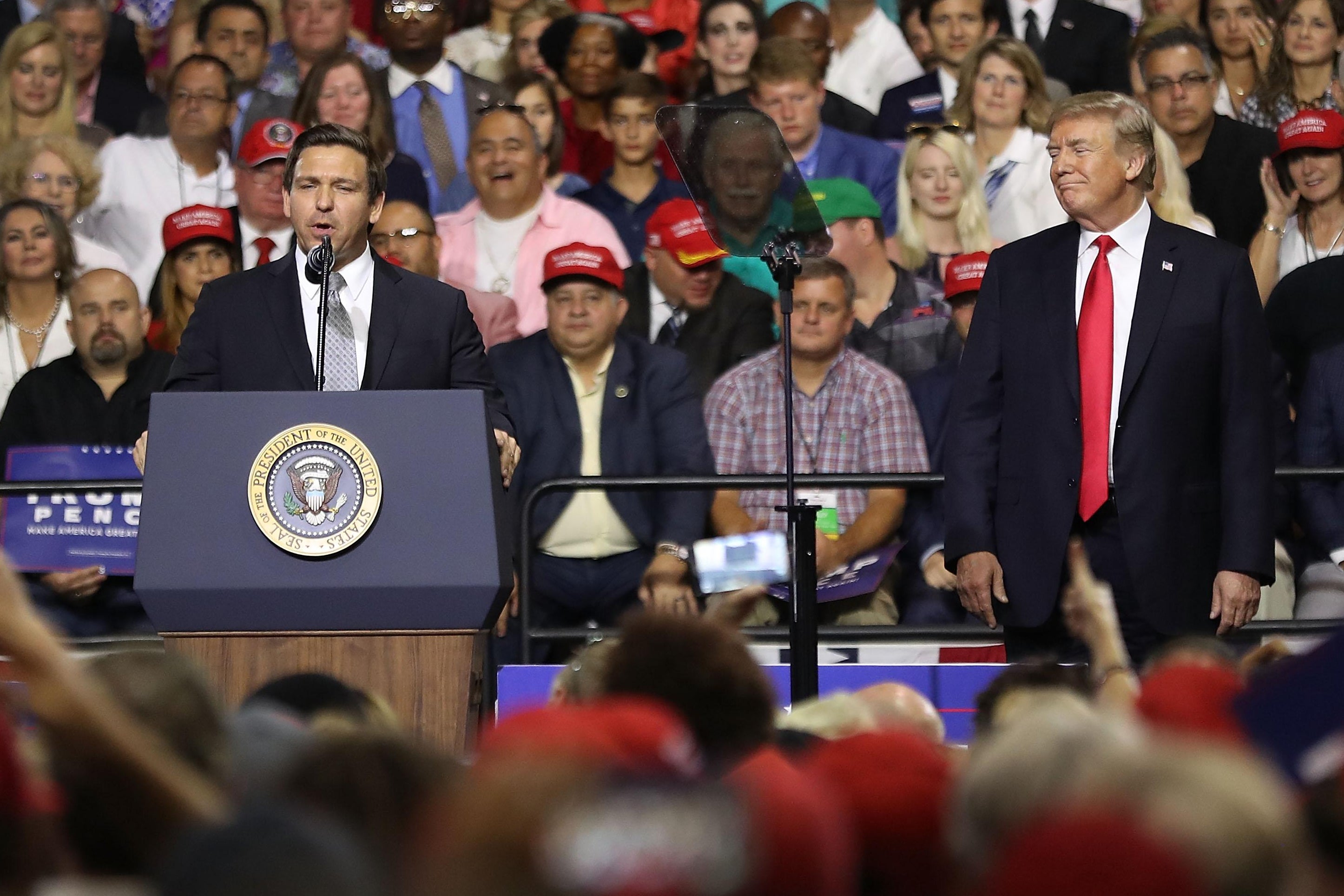 Ron DeSantis speaking at a lectern while Donald Trump watches from the side of the stage