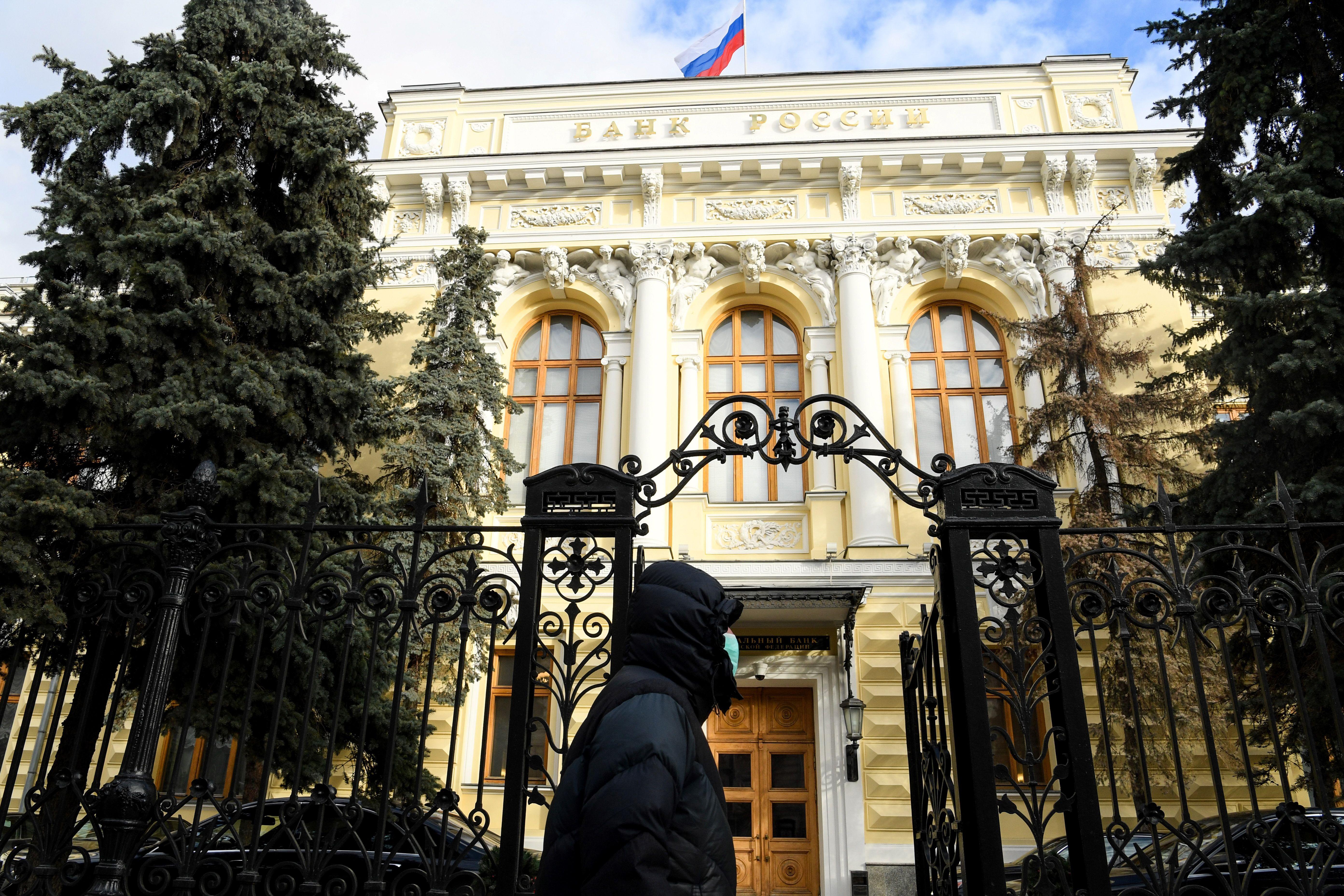 A man in a face mask in front of an iron gate and a large building flying a Russian flag.