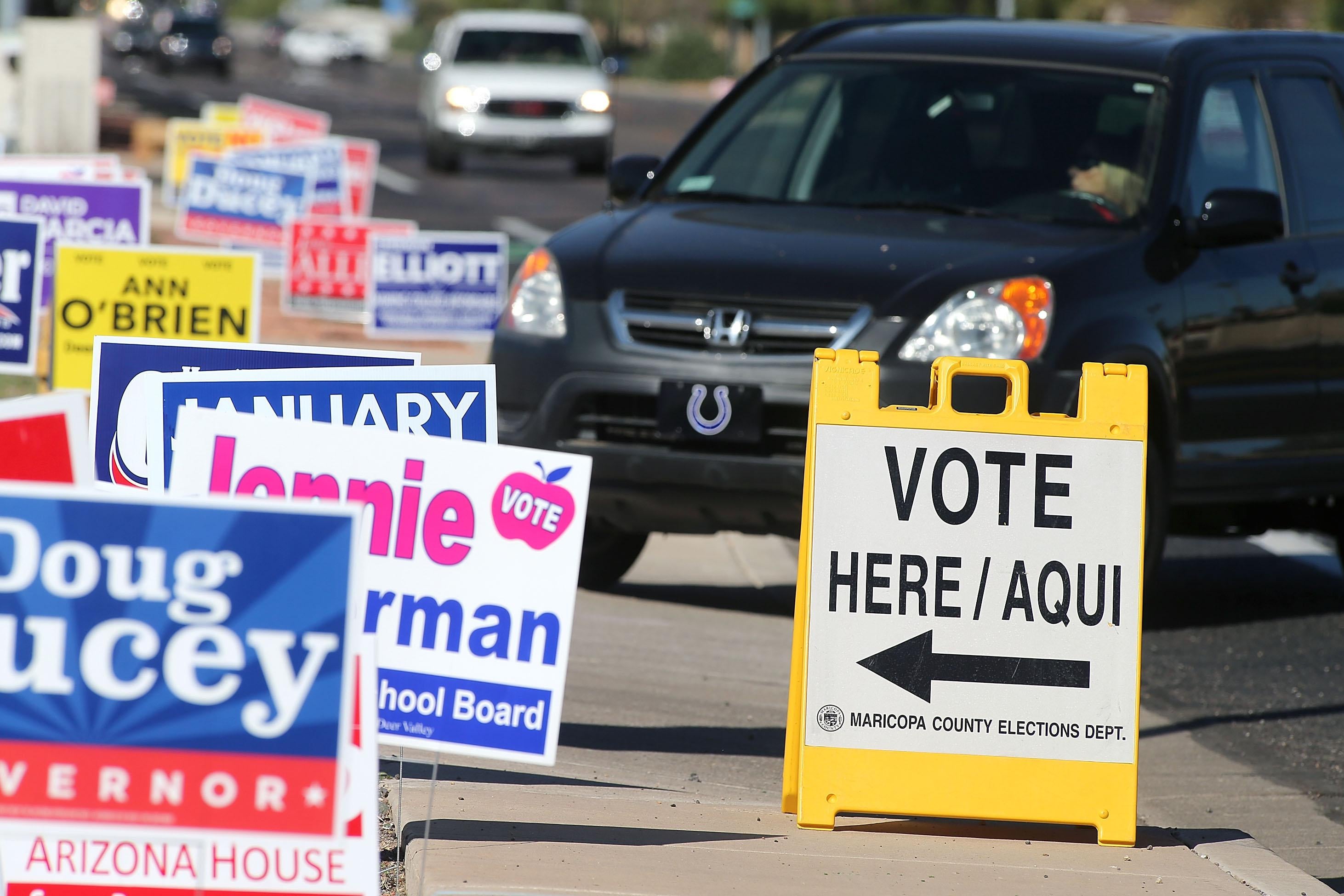 A car drives by a "VOTE HERE/AQUI" sign and a row of campaign signs for Doug Ducey and others.