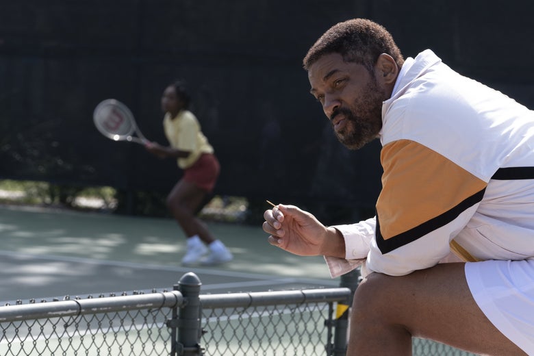 A girl in a yellow shirt and red shorts is in the background playing tennis while, in the foreground, her father chews a toothpick; he wears a white track suit.