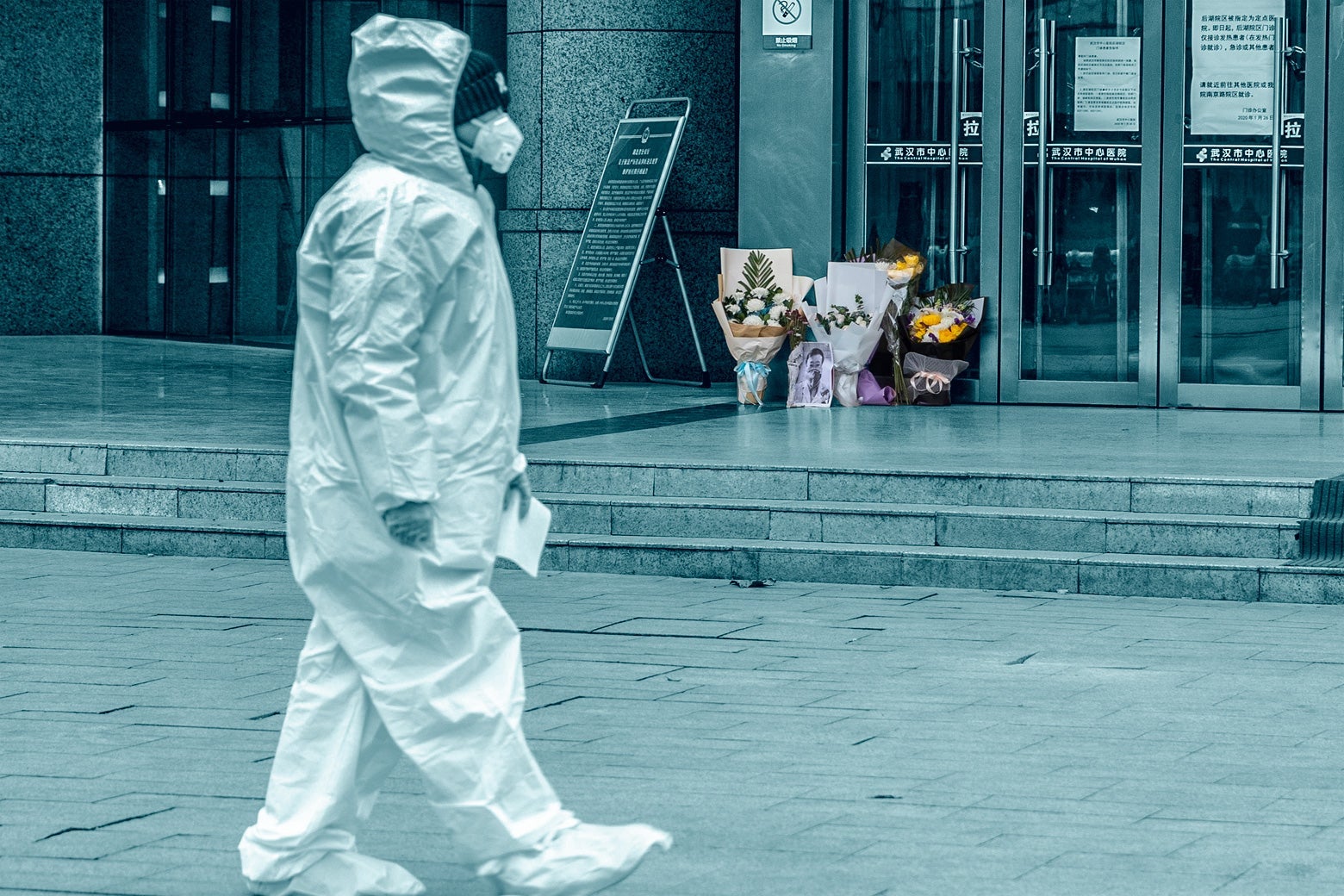 A medical staff member in full-body protective gear walks past a flower tribute on the ground outside the hospital doors