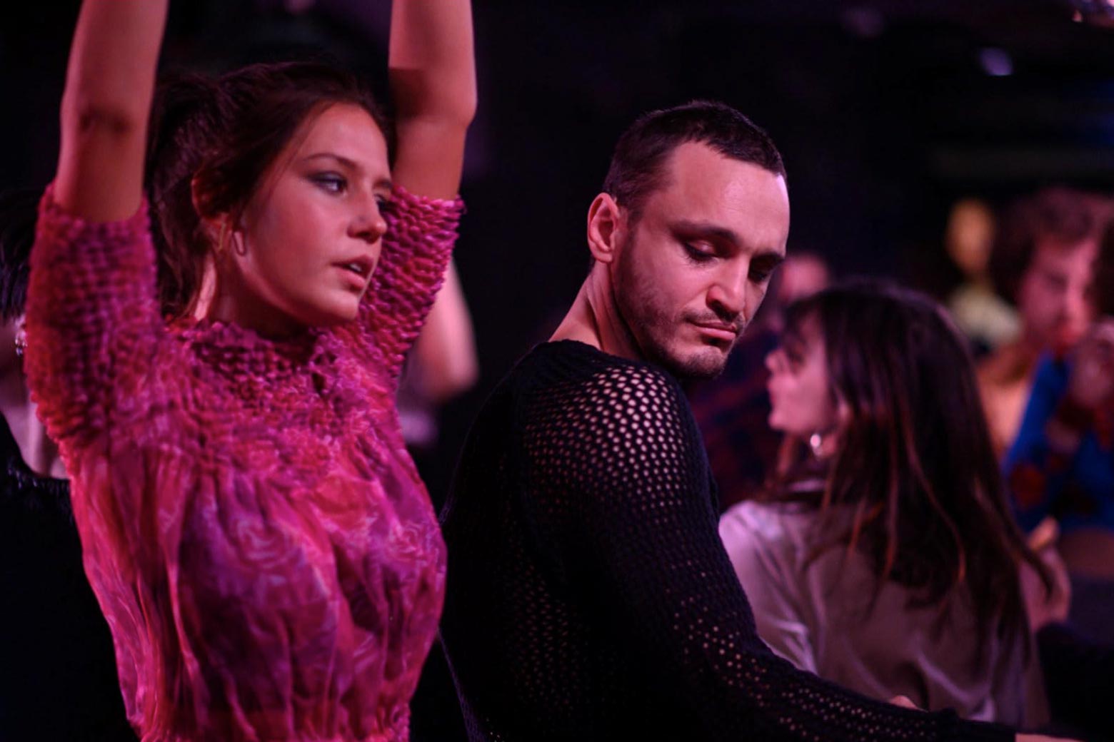 In the dim light of a club, a woman wearing a fuchsia top puts her hands up in the air and dances, while a man standing next to her glances seductively in her direction.