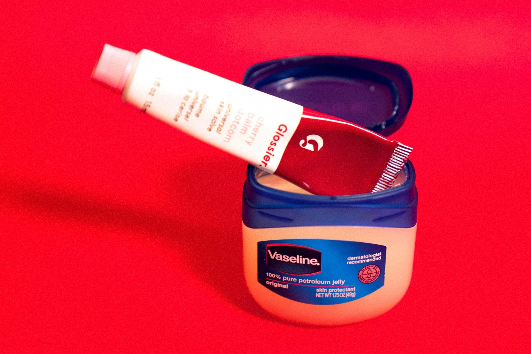 A tube of Glossier Balm Dotcom perched on top of an open jar of Vaseline.