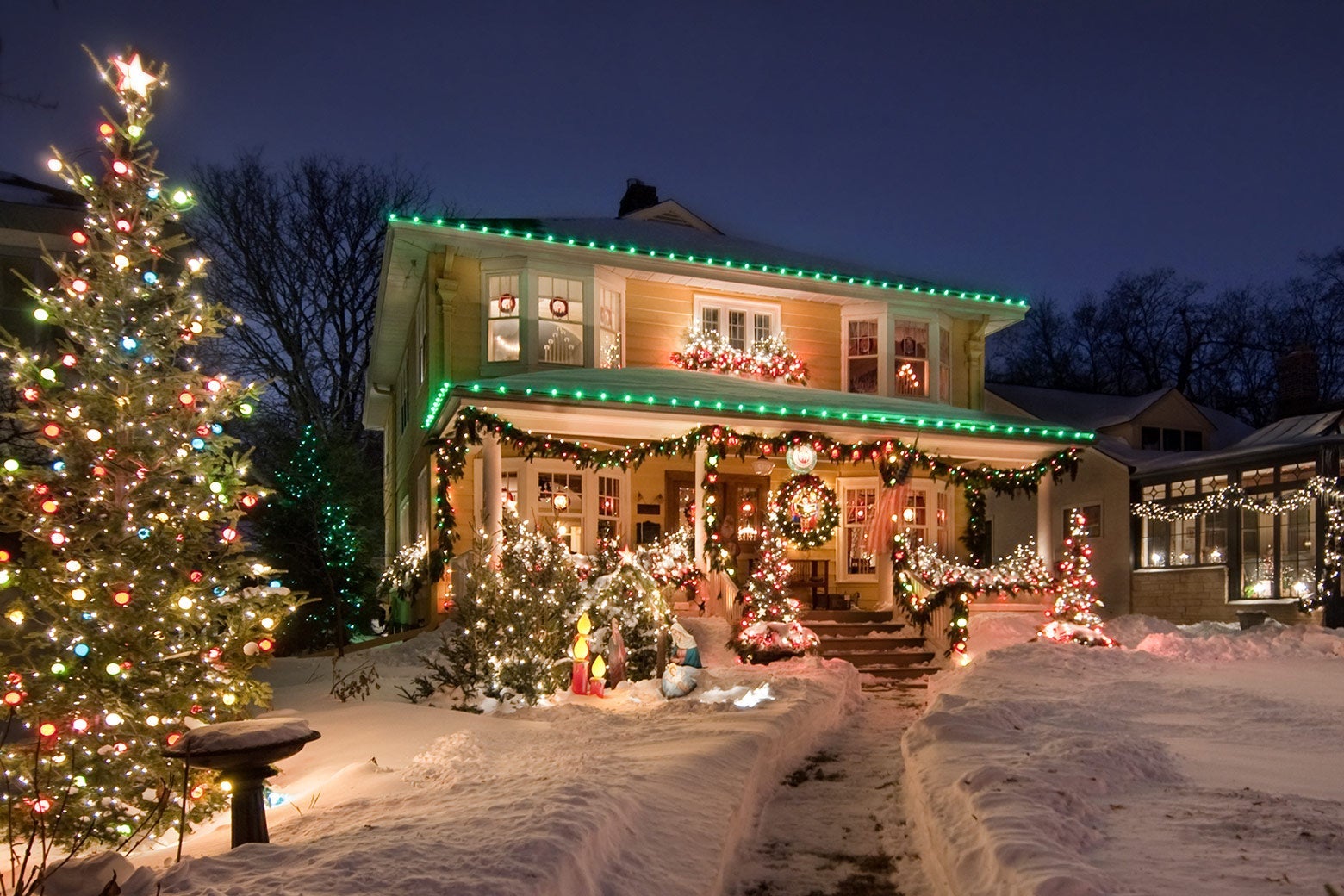 A house with an elaborate holiday light display out front, with multiple Christmas trees on a snow-covered front lawn