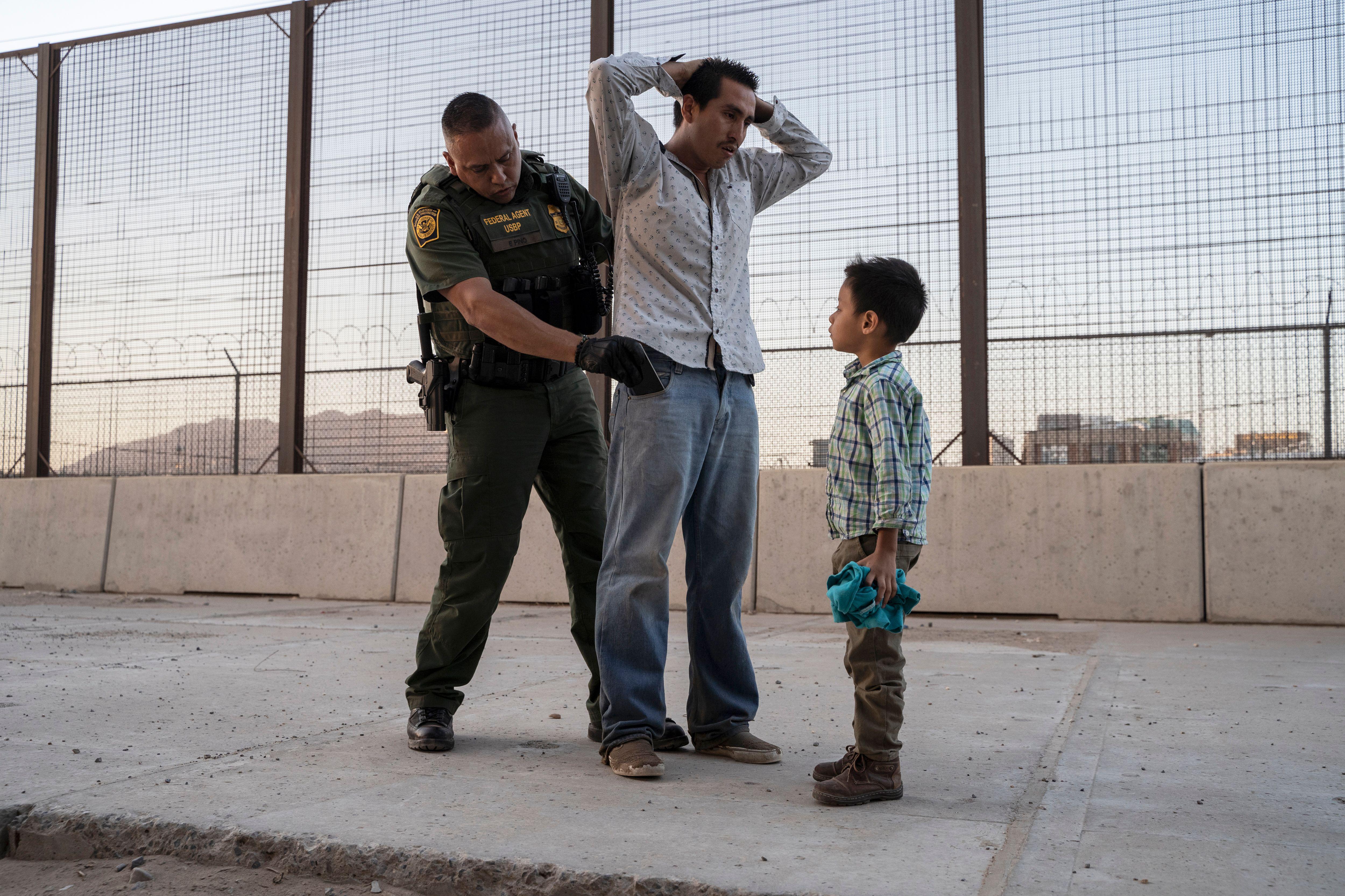A migrant father has his hands clasped behind his head as a CBP agent searches his pockets. The migrant's son looks on.