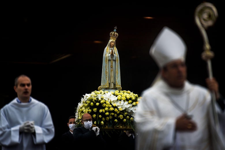 A statue of Mary praying on a bed of flowers is held aloft by men in face masks. Two people in Catholic vestments—the one in the foreground a bishop—are out of focus, walking with the statue.