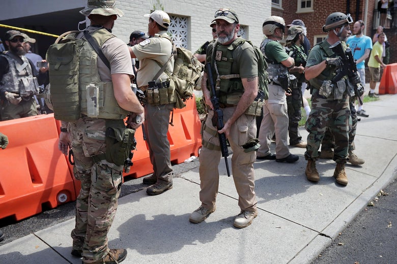 Several men wearing body armor and carrying guns stand on the sidewalk.