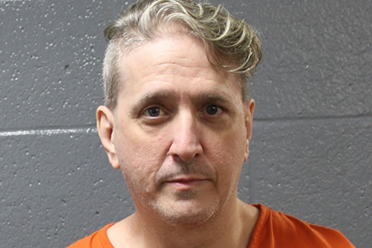 A man in an orange jumpsuit with wavy blonde hair and a thin mustache.