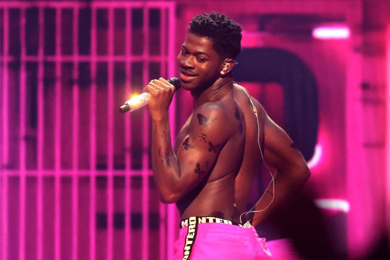 A shirtless man wears bright pink pants with a belt that says MONTERO. Behind him are pink jail cell bars. He smiles and holds a gold microphone up to his mouth.