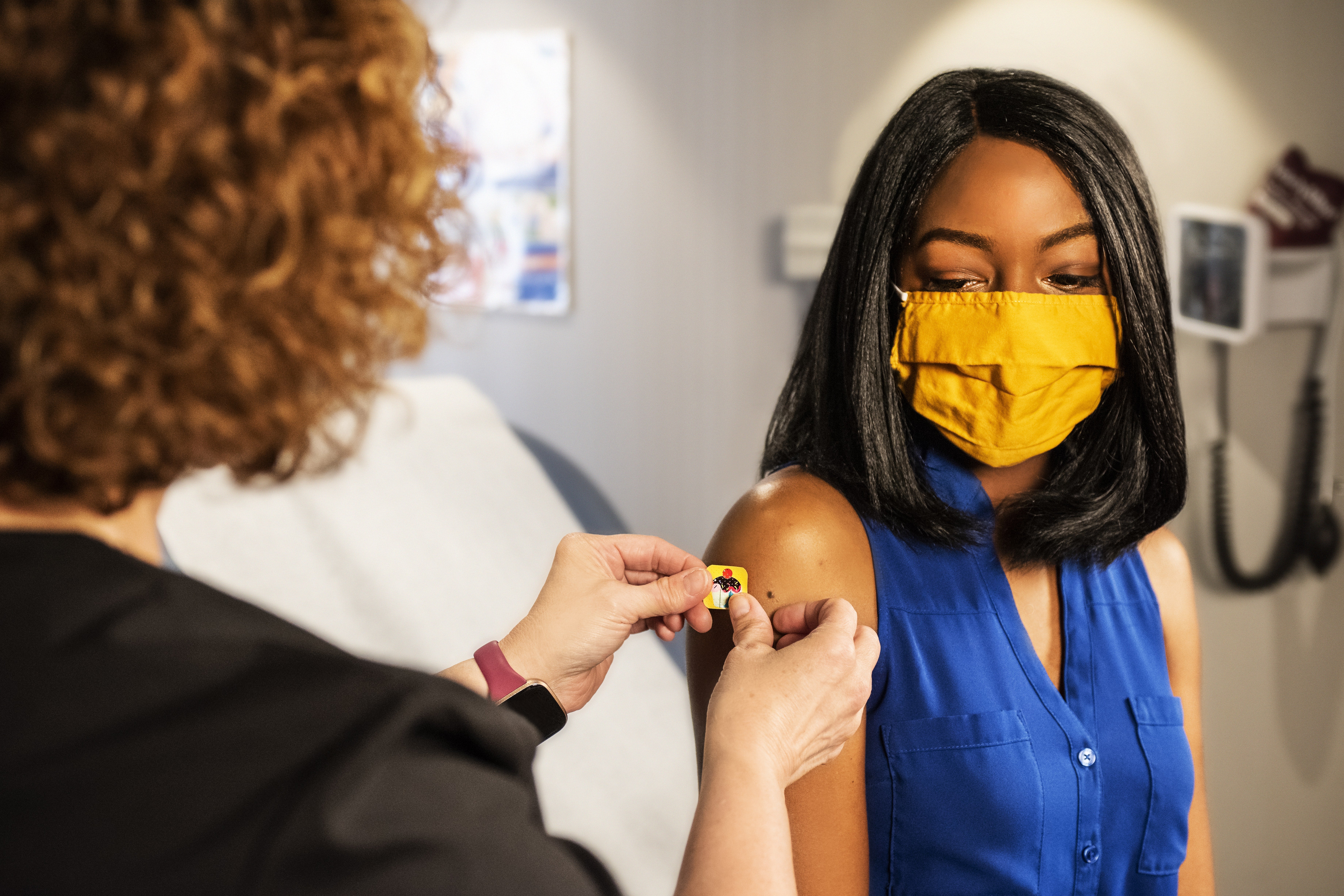 A medical worker puts a band-aid on the upper arm of a woman wearing a mask in an exam room