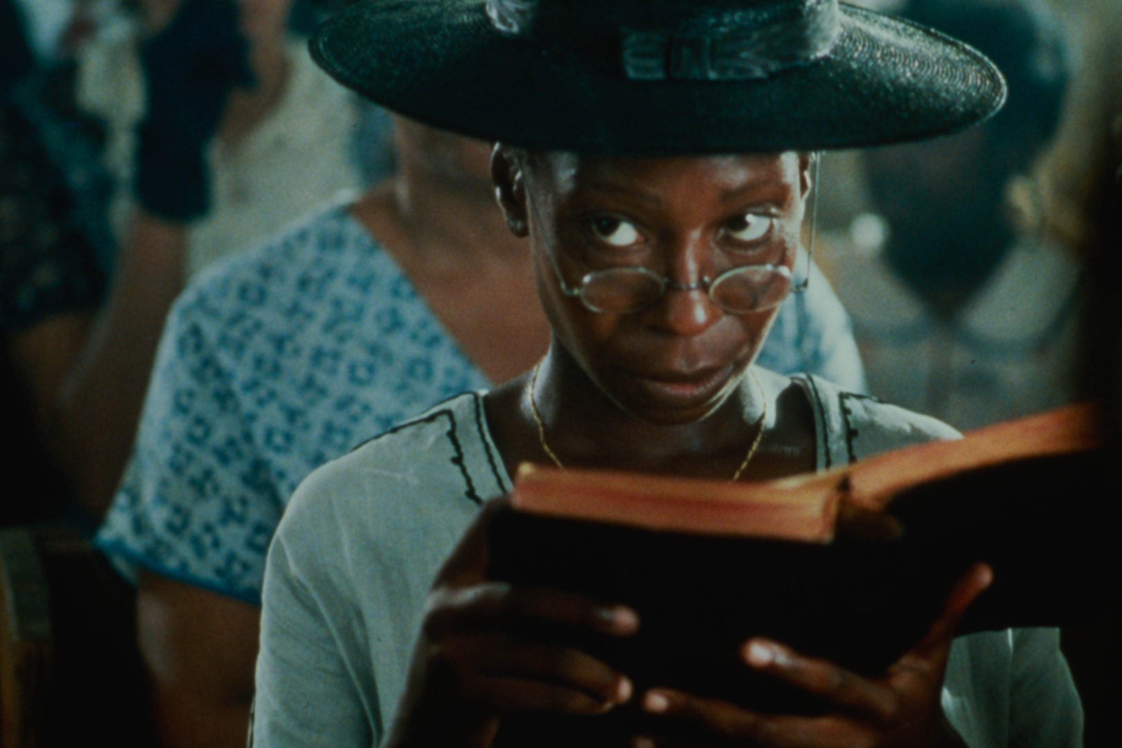A black woman in a large black hat sits in a church pew looking over her glasses at something off-screen while holding a bible in her hands.