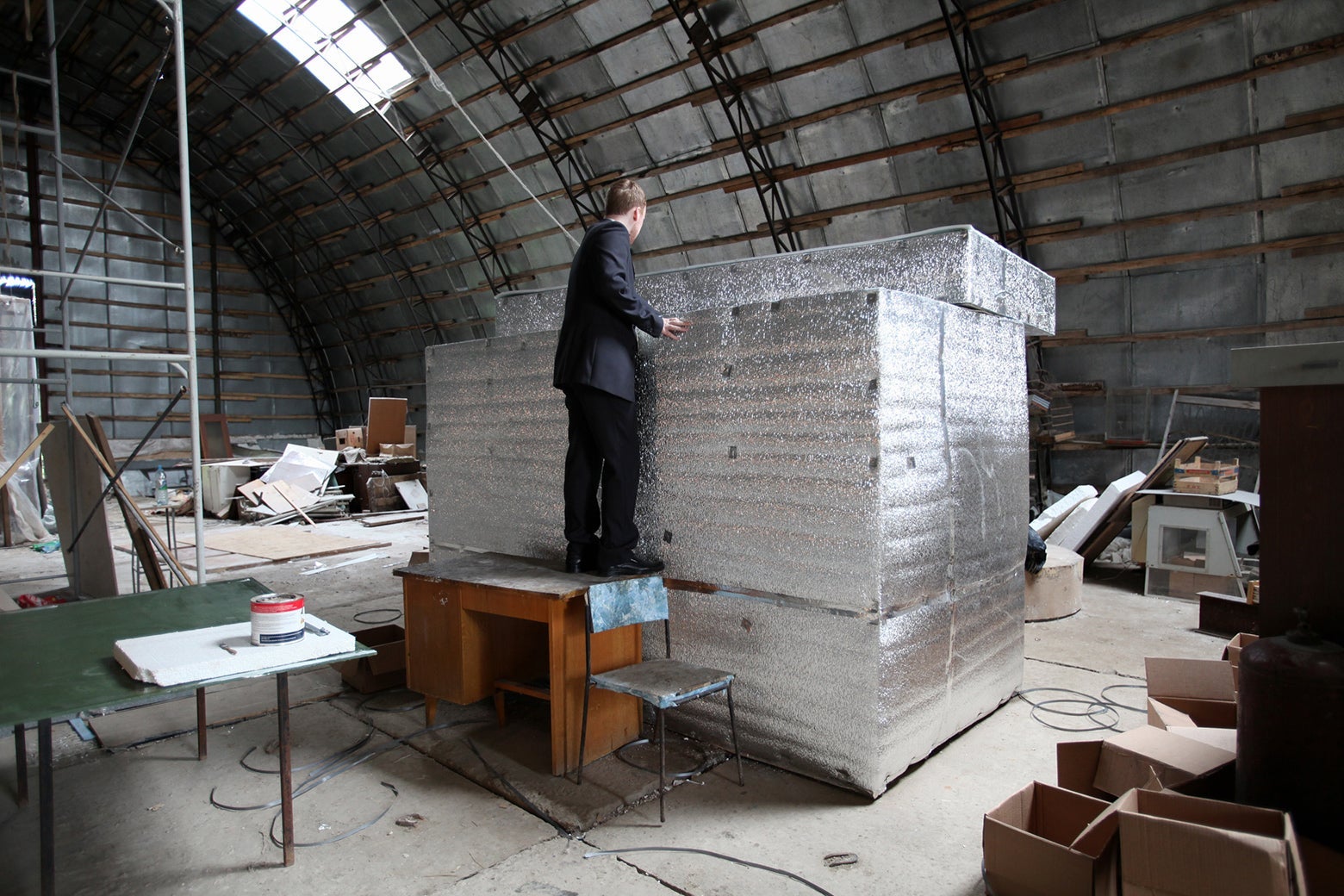 In the center of a cluttered warehouse room, a man stands on a desk to look inside a large silver container.