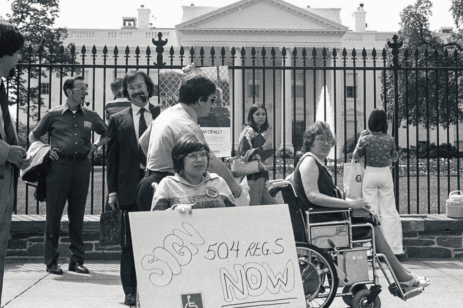 A black-and-white photo of protestors, some of them in wheelchairs, rallying outside of the White House. One of the signs encourages the president to "Sign 504 now."