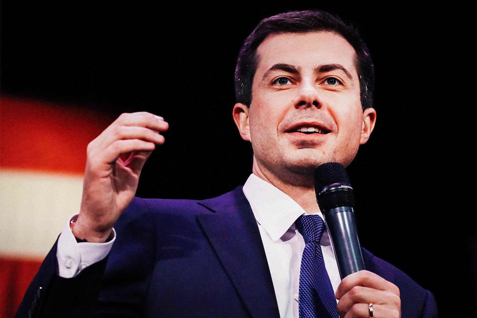 Pete Buttigieg holding a mic and speaking with an American flag in the background