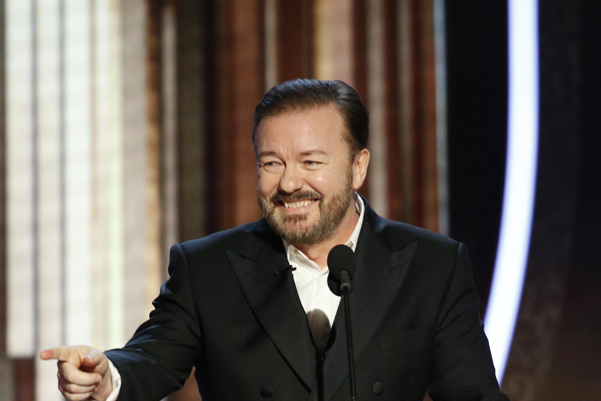 Gervais, standing at a podium, grins and points with his right hand.