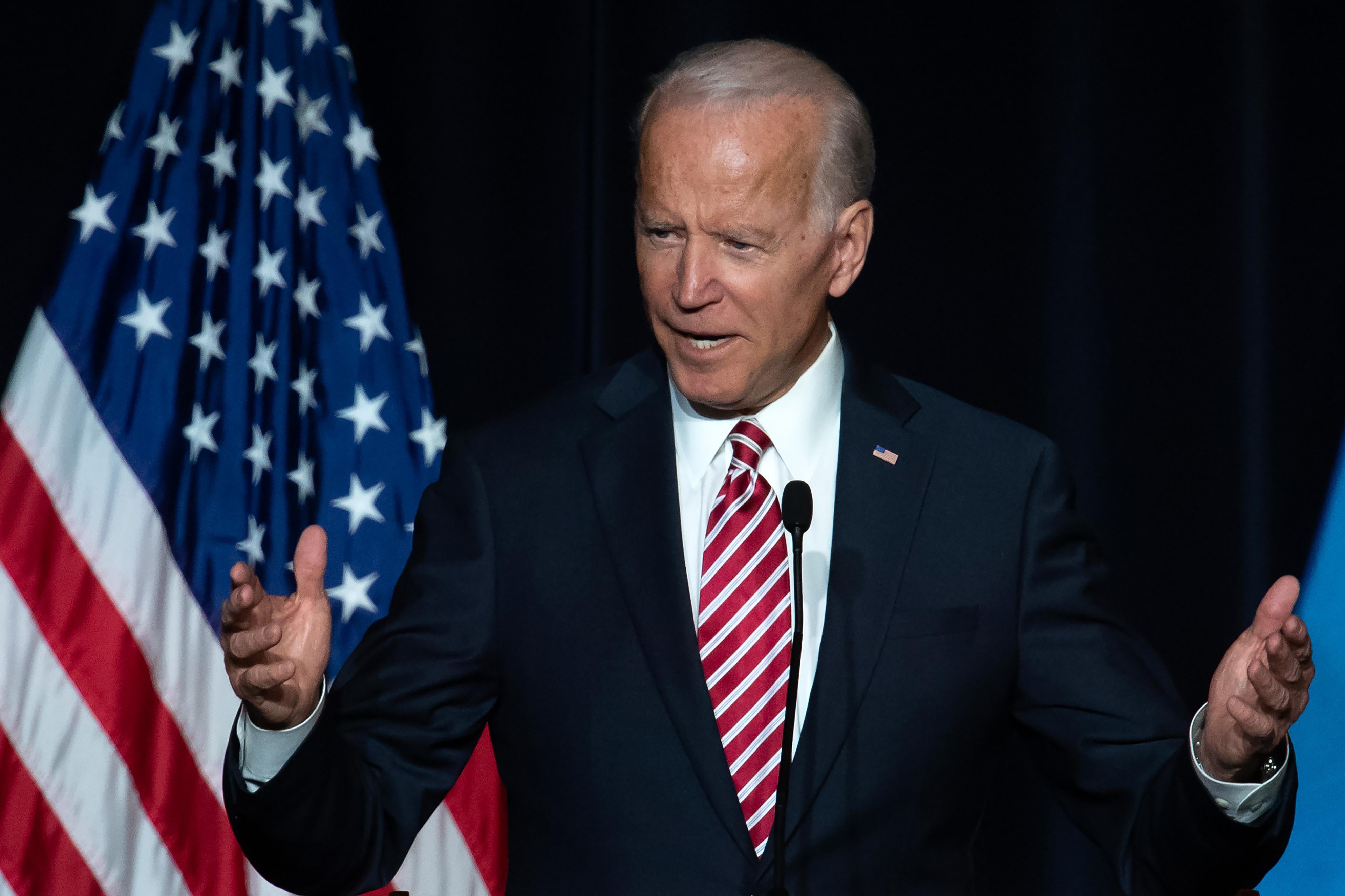 Biden speaking at a mic, arms outstretched, with an American flag behind him.