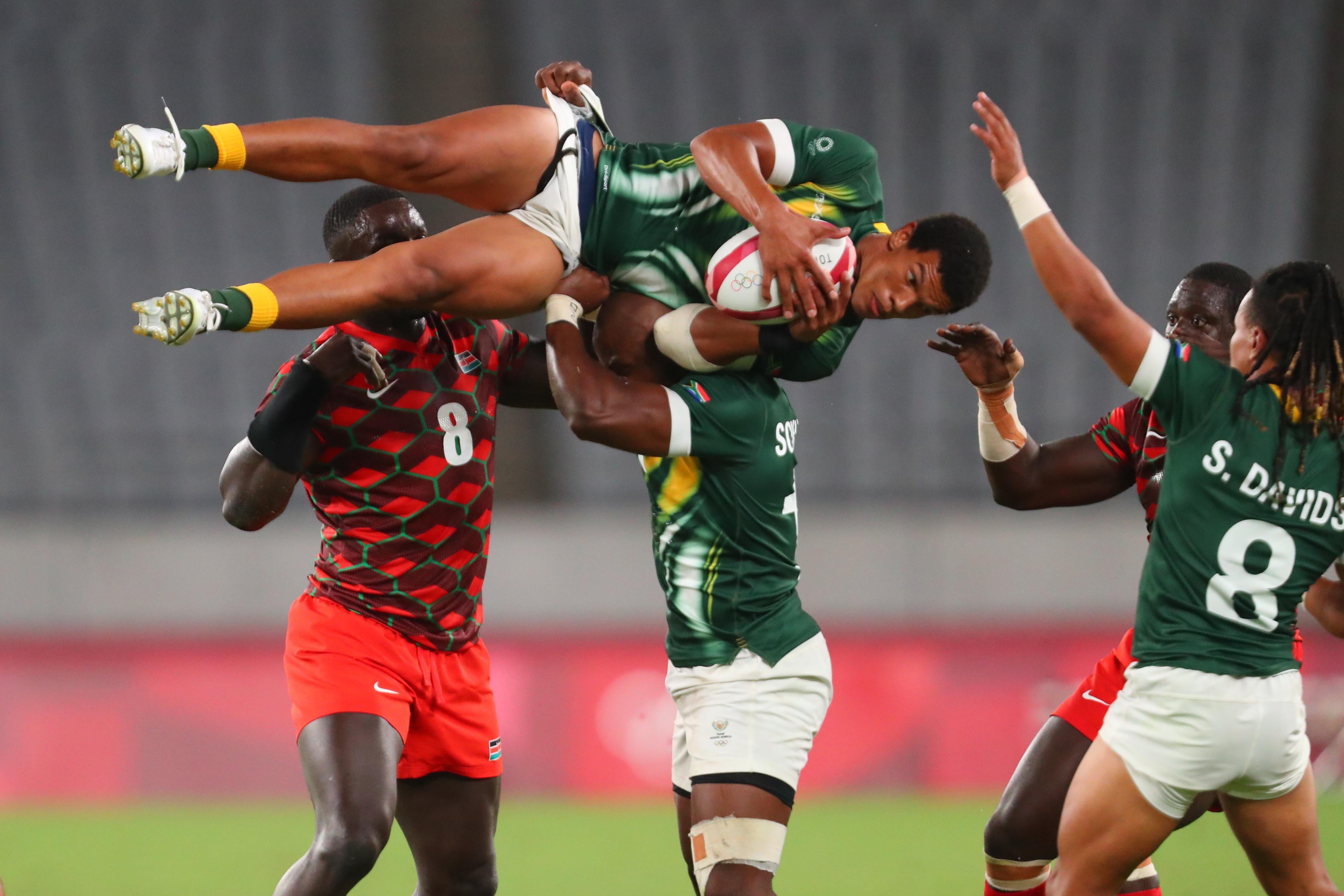 A rugby player in a green and white uniform holding the ball, is hoisted up horizontally by another player, as several other rugby players stand around him
