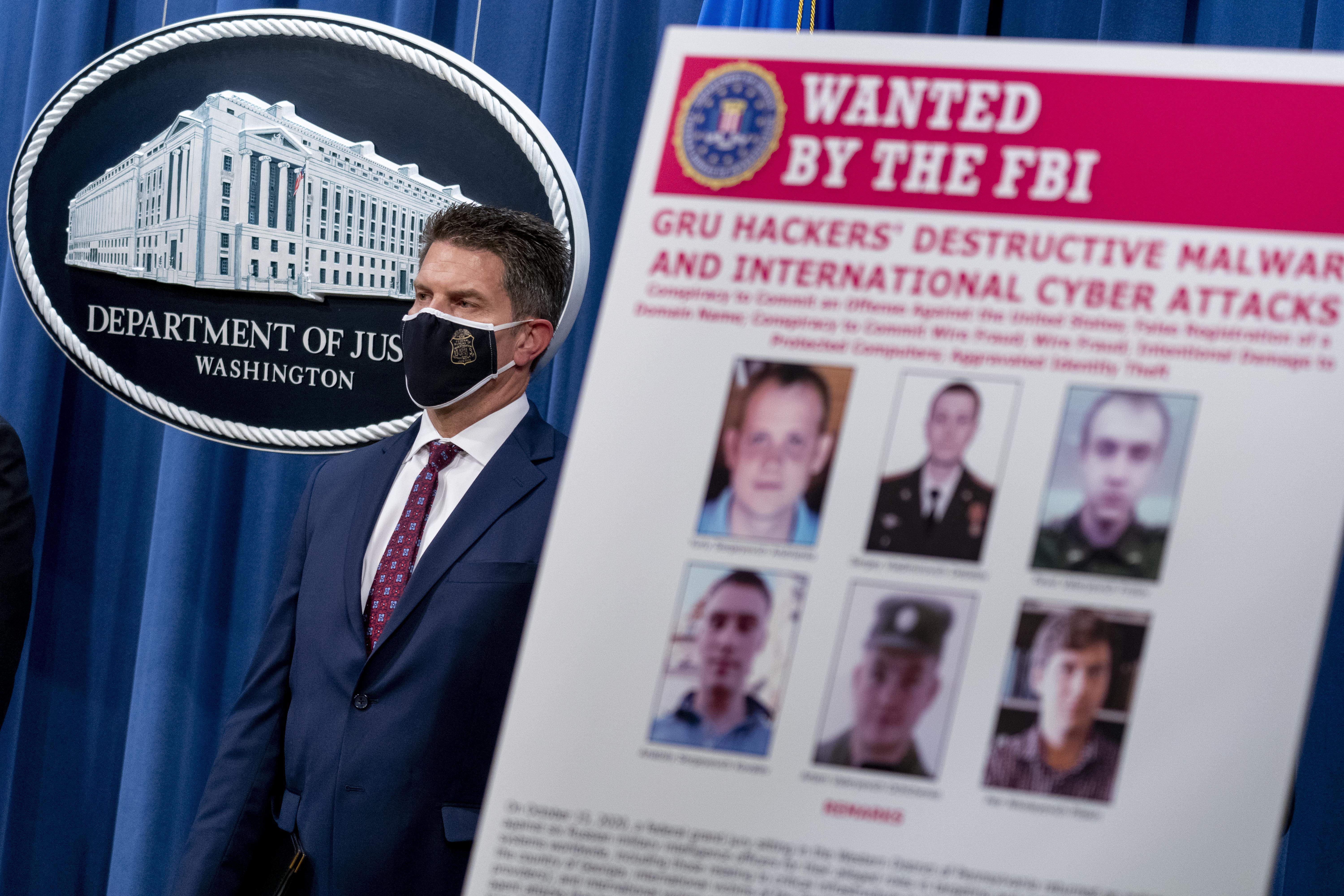 A poster with six photos says "Wanted by the FBI" in front of a man wearing a mask next to the seal of the Department of Justice.