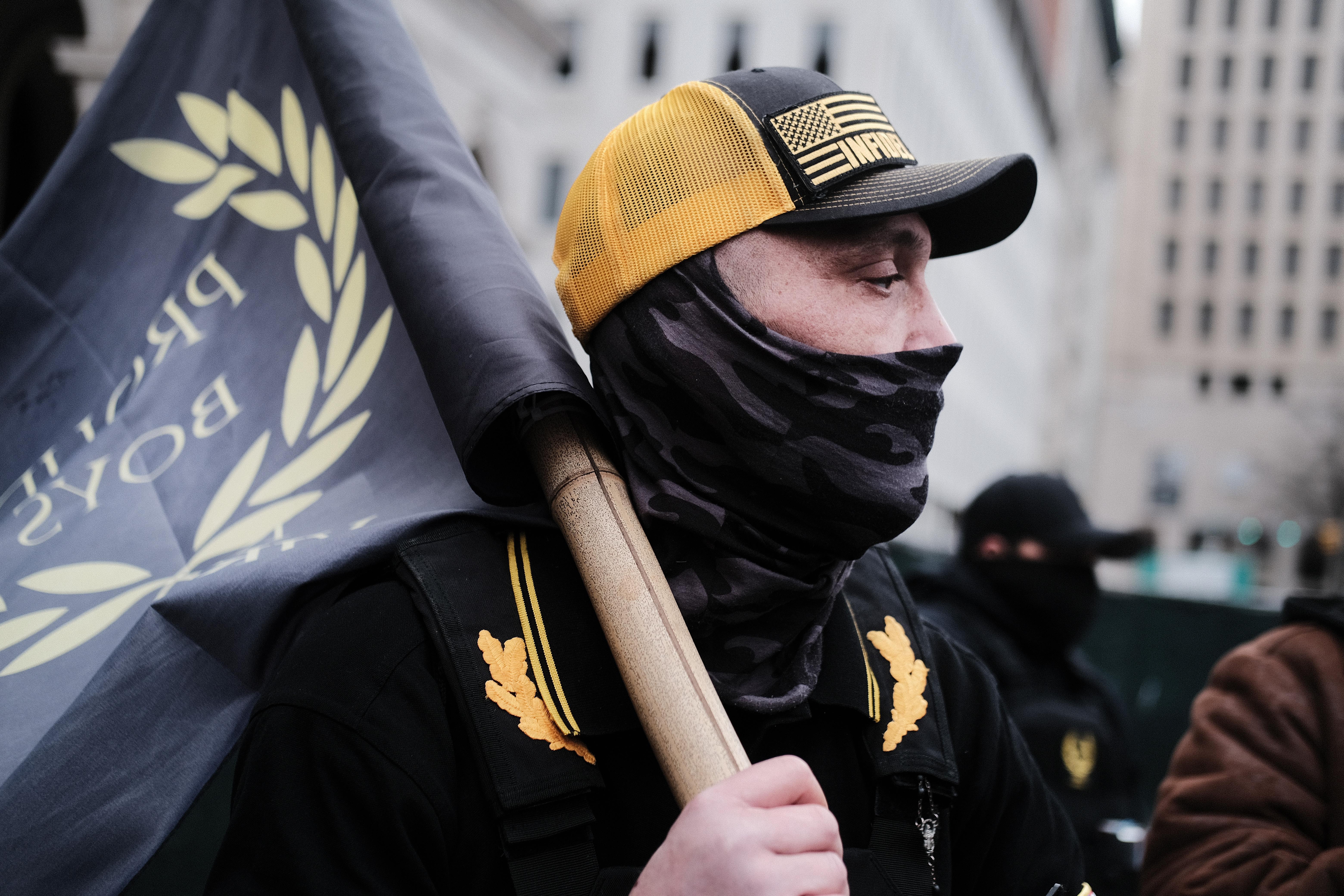 A man in black and yellow clothing carrying a Proud Boys flag. 