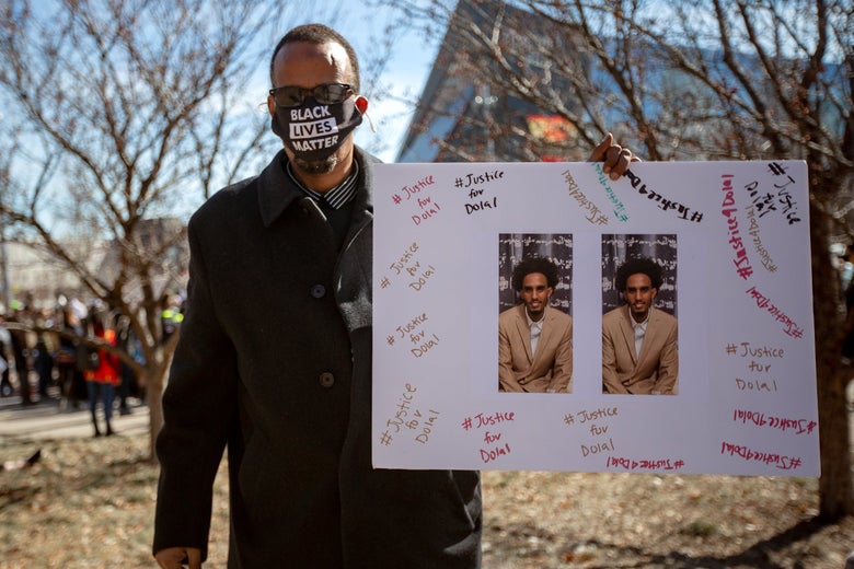 A man wearing a Black Lives Matter mask holds up a poster with photos of Dolal Idd surrounded by the words "Justice for Dolal"