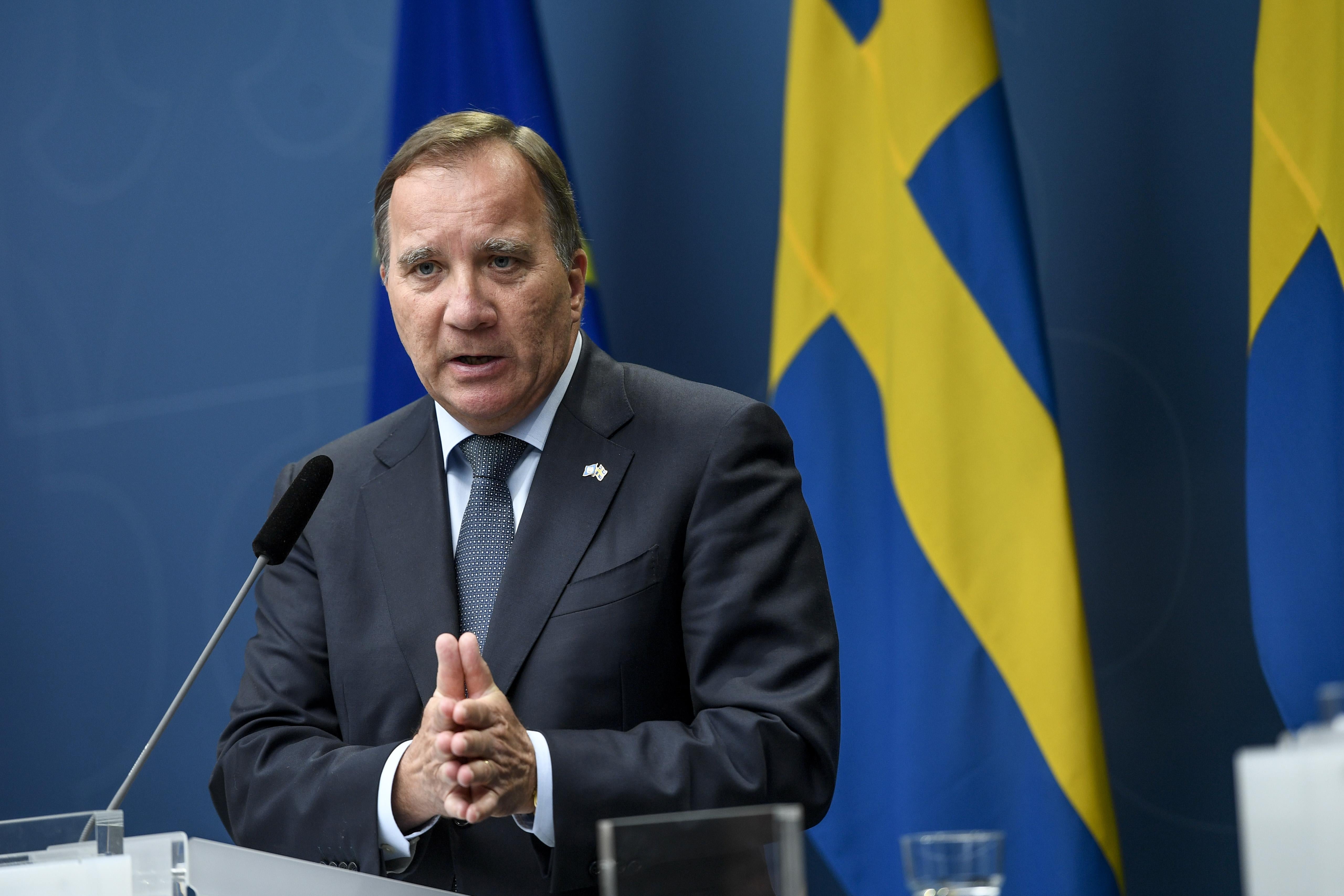 Stefan Löfven stands in front of a Swedish flag and speaks into a microphone while folding his hands.