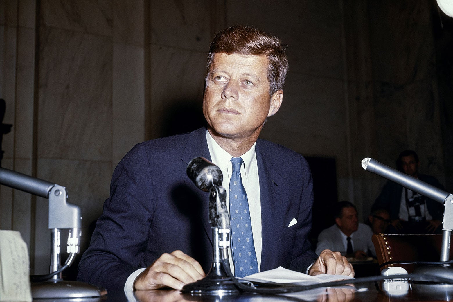 John F. Kennedy in front of a bank of microphones on a desk.