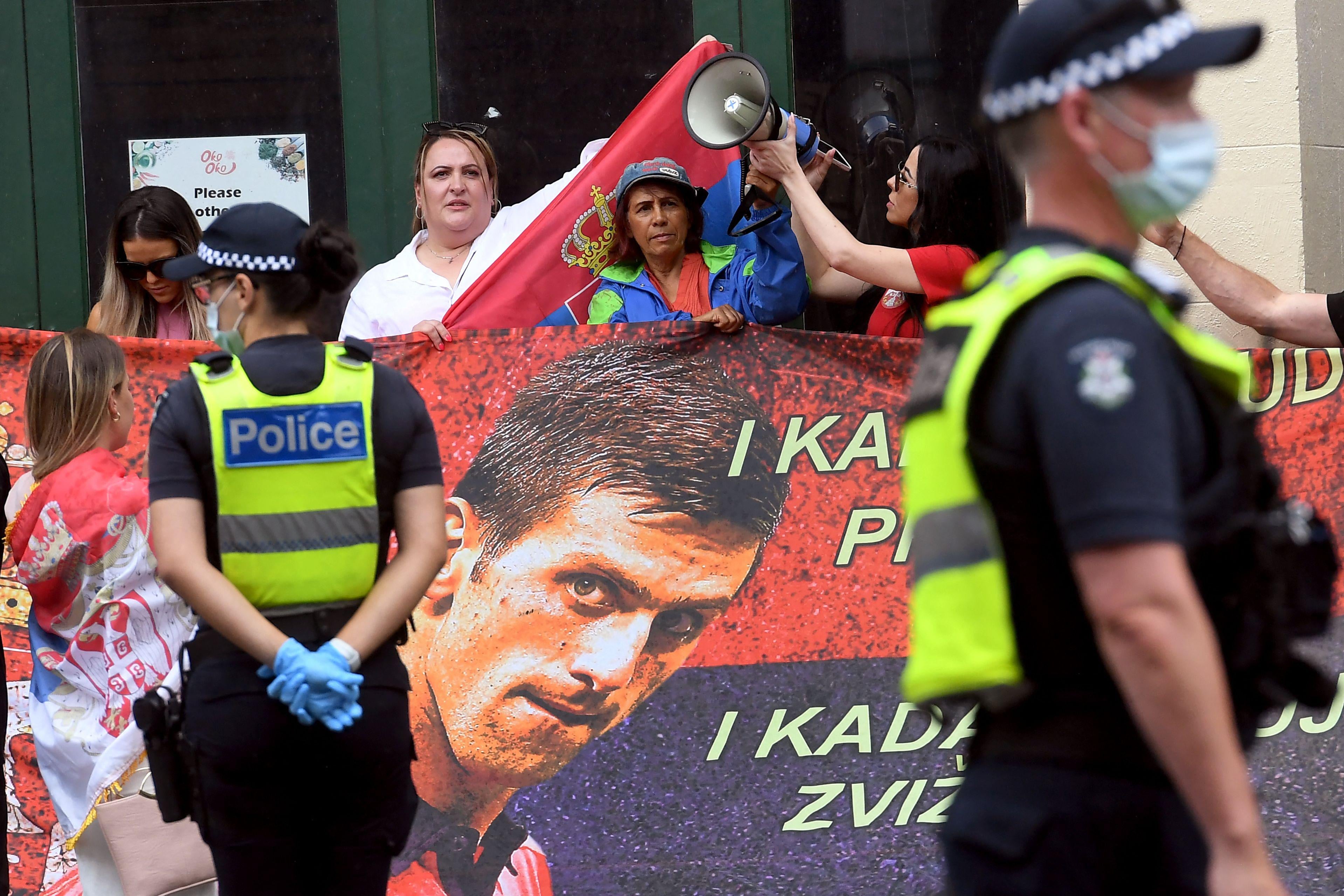 Novak supporters hold sings of support, a big one showing Djokovic's face, as police patrol the scene