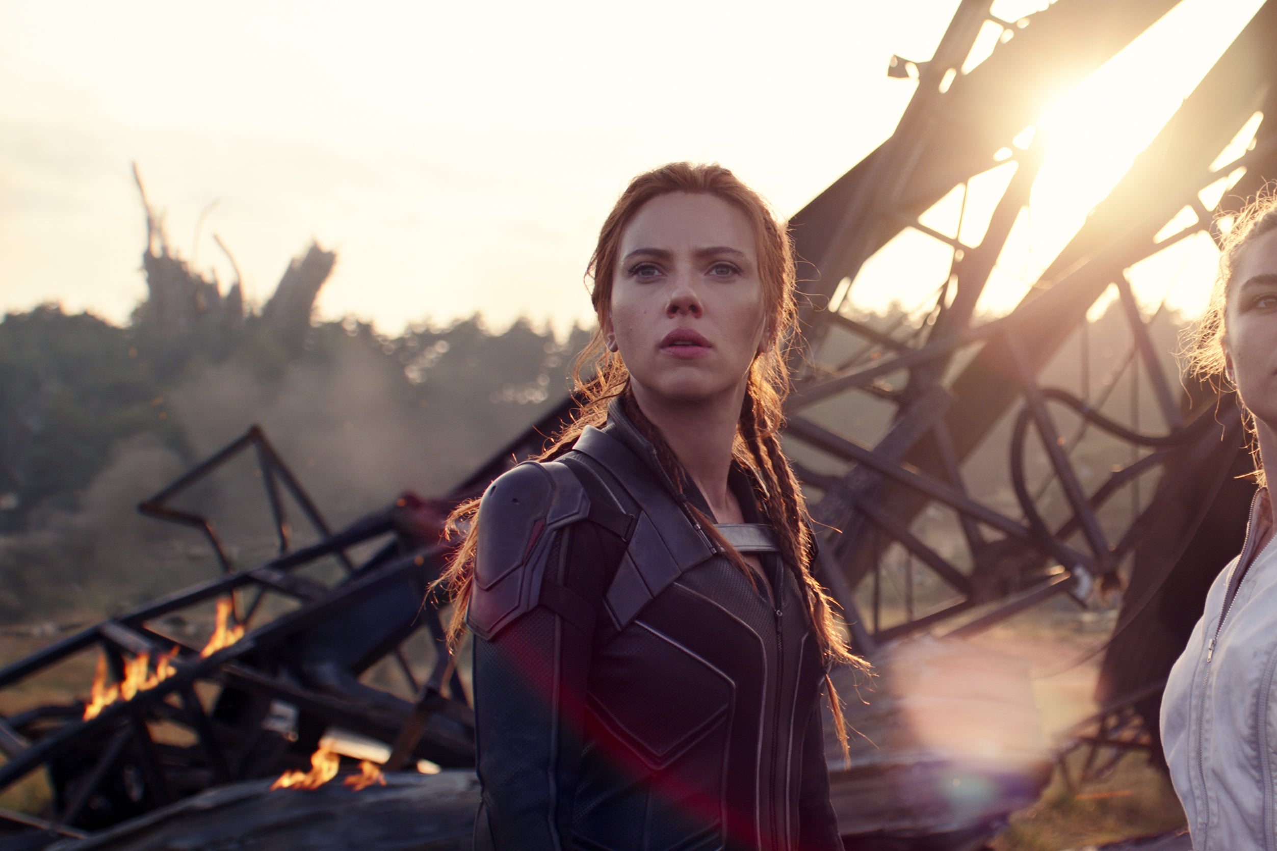 The two women stand in supersuits in front of wreckage, their hair tied back.