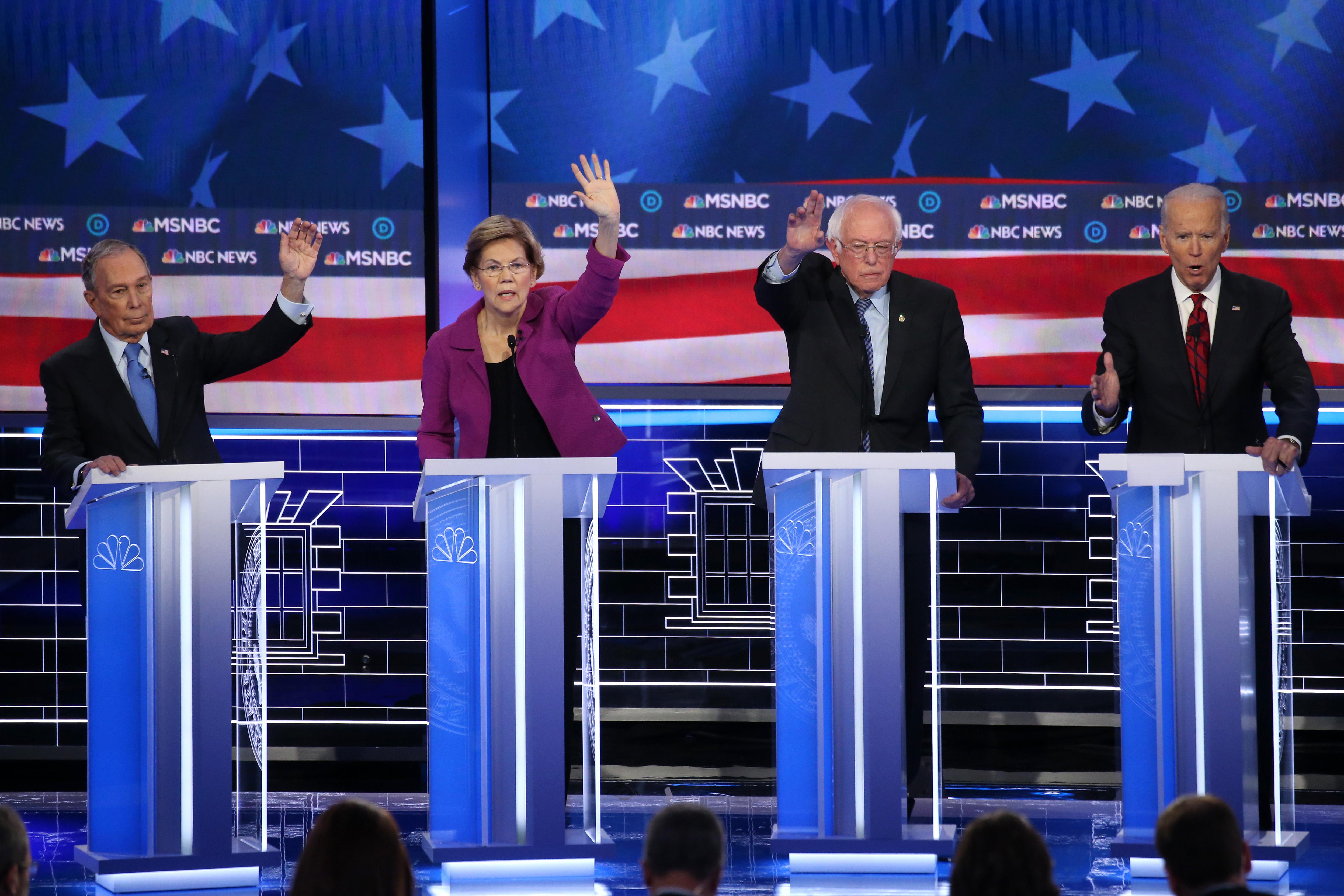 Michael Bloomberg, Elizabeth Warren, and Bernie Sanders raise their hands on the debate stage as Joe Biden speaks.