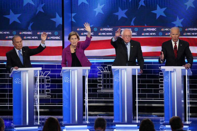 Michael Bloomberg, Elizabeth Warren, and Bernie Sanders raise their hands on the debate stage as Joe Biden speaks.