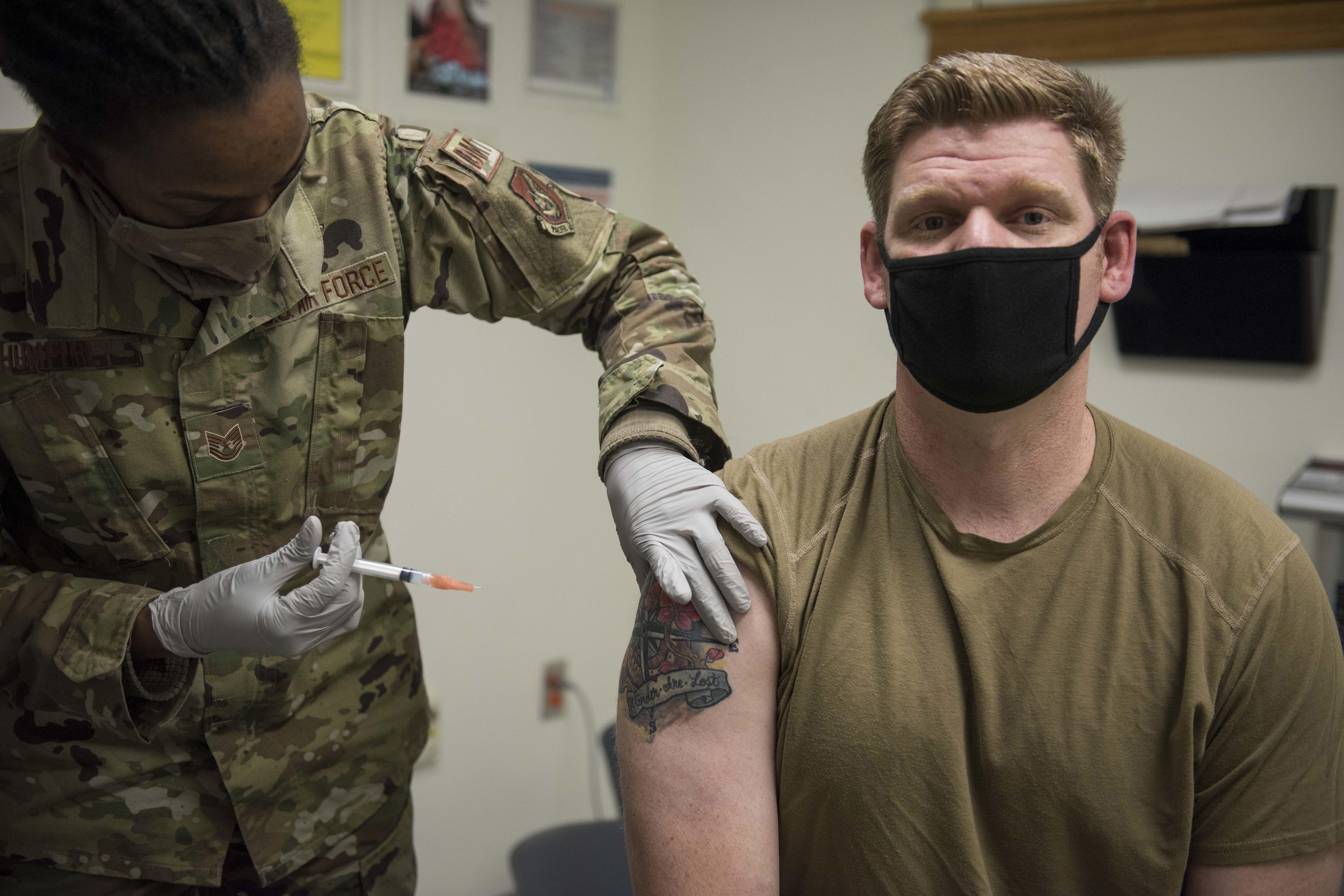 A man in military-green T-shirt and face mask looks at the camera as a woman in fatigues holds a needle near his bicep.