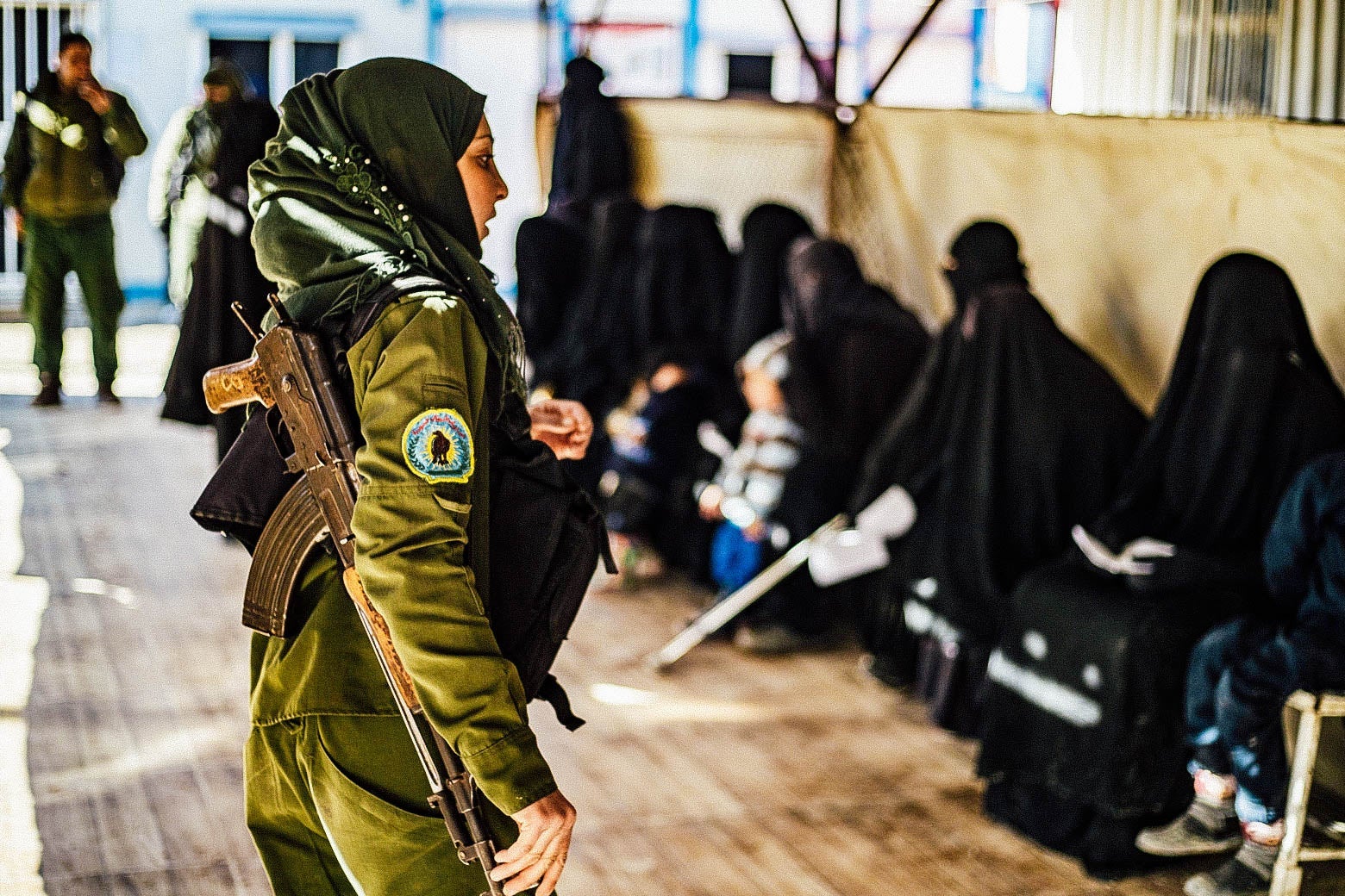 A female Kurdish fighter in uniform faces a row of women seated, wearing black veils.