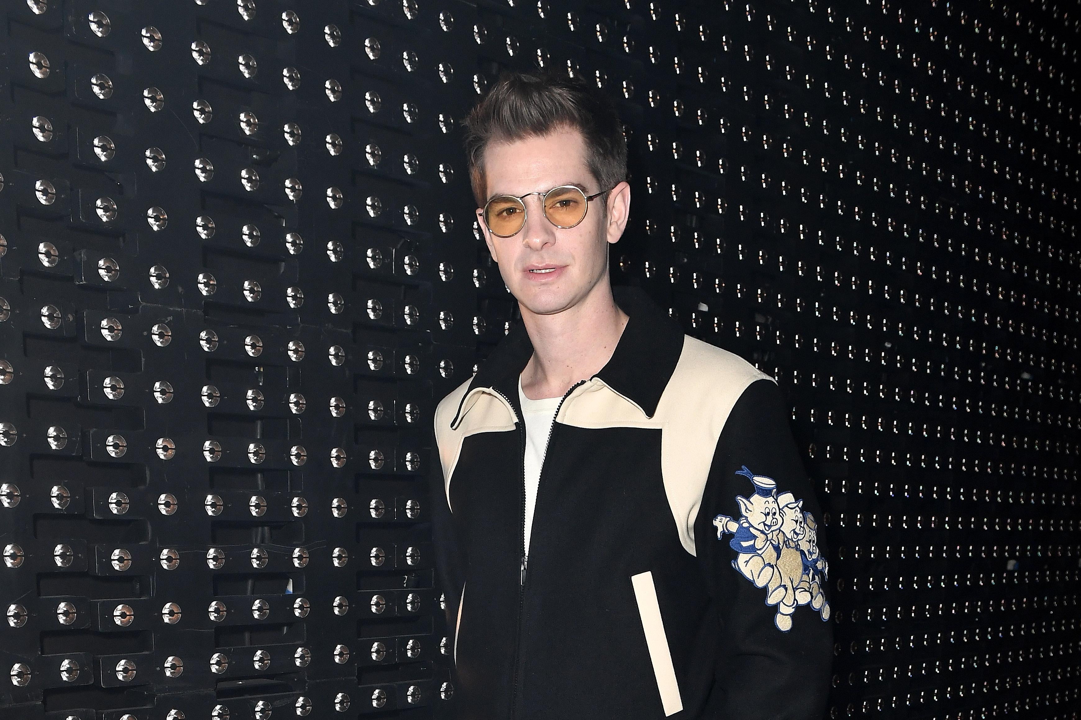 Andrew Garfield, wearing sunglasses and a jacket with cartoon pigs on the sleeve, poses in front of a black studded wall.