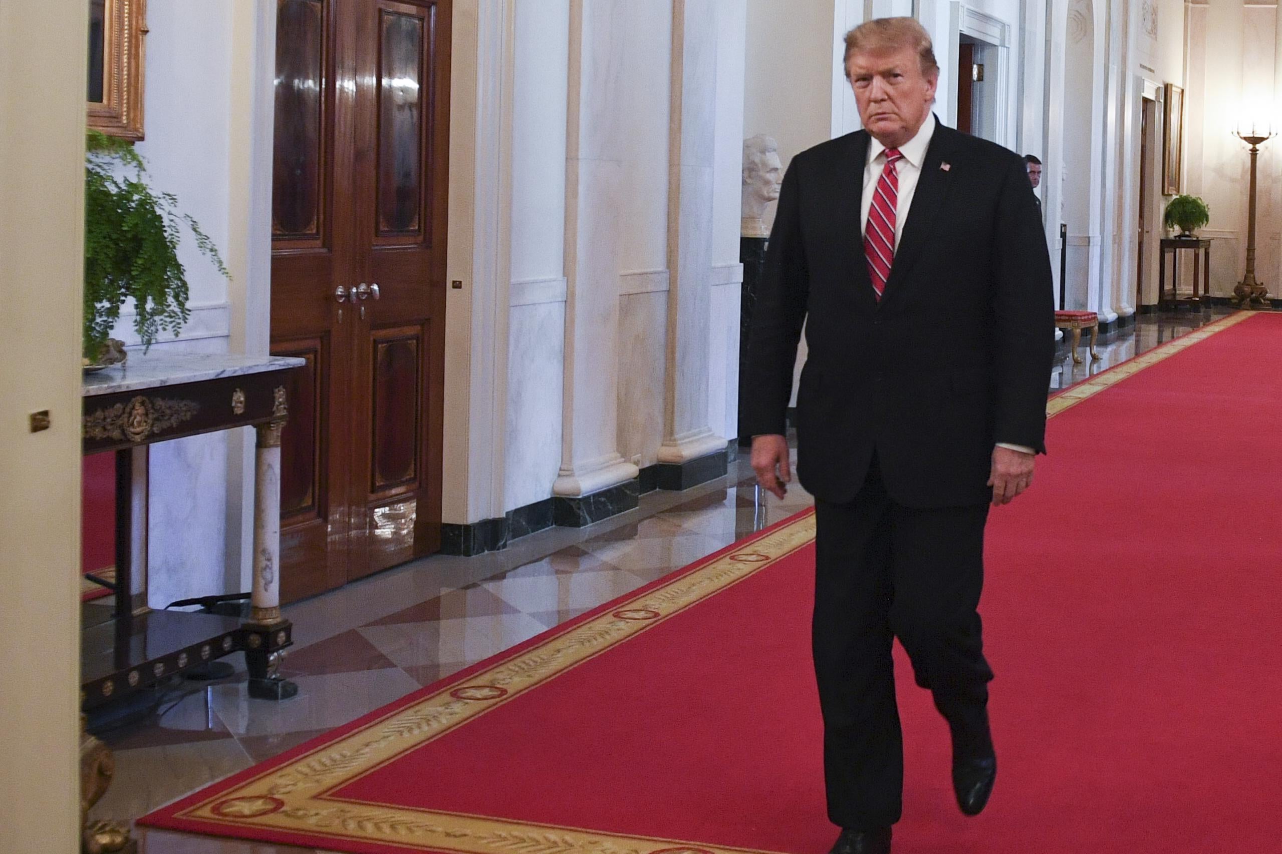 Trump walking down a red-carpeted hallway in the White House.