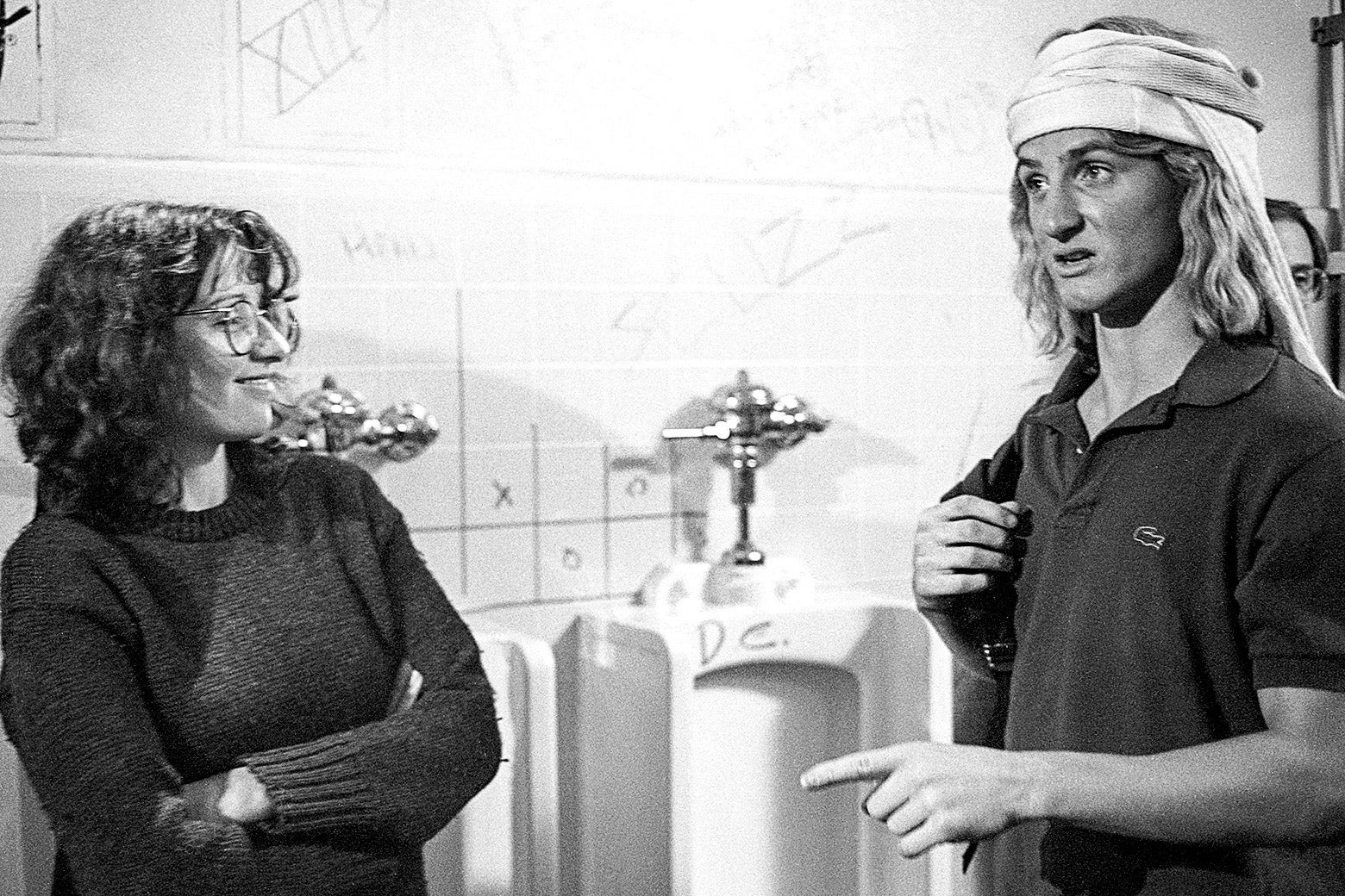 Black and white photo of Heckerling smiling with arms crossed at Penn as Spicoli wearing a polo and backpack in a boys bathroom with a row of urinals behind them