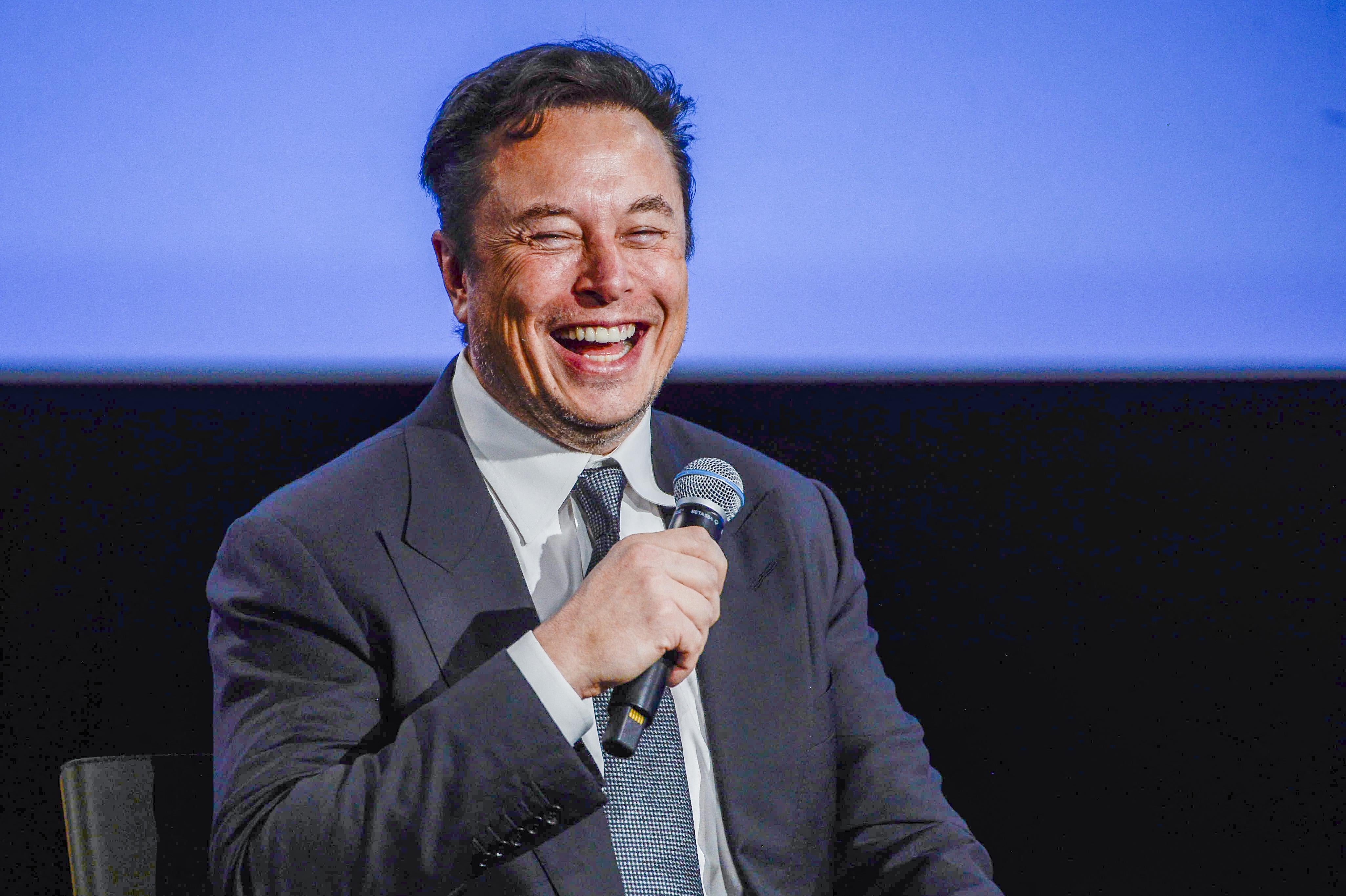 A seated Musk, wearing a suit, grins as he holds a mic on a stage.